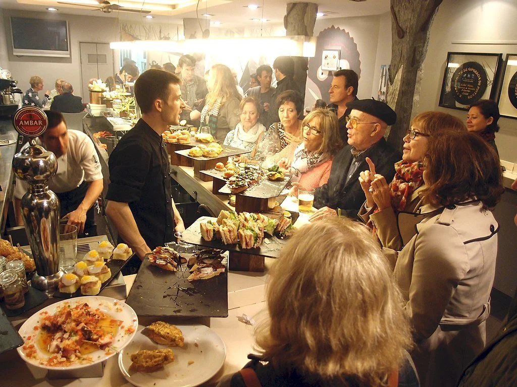 Diners eat a late dinner at a bar crowded with food and drinks in San Sebastian, Spain, while talking to a man in black shirt behind the counter