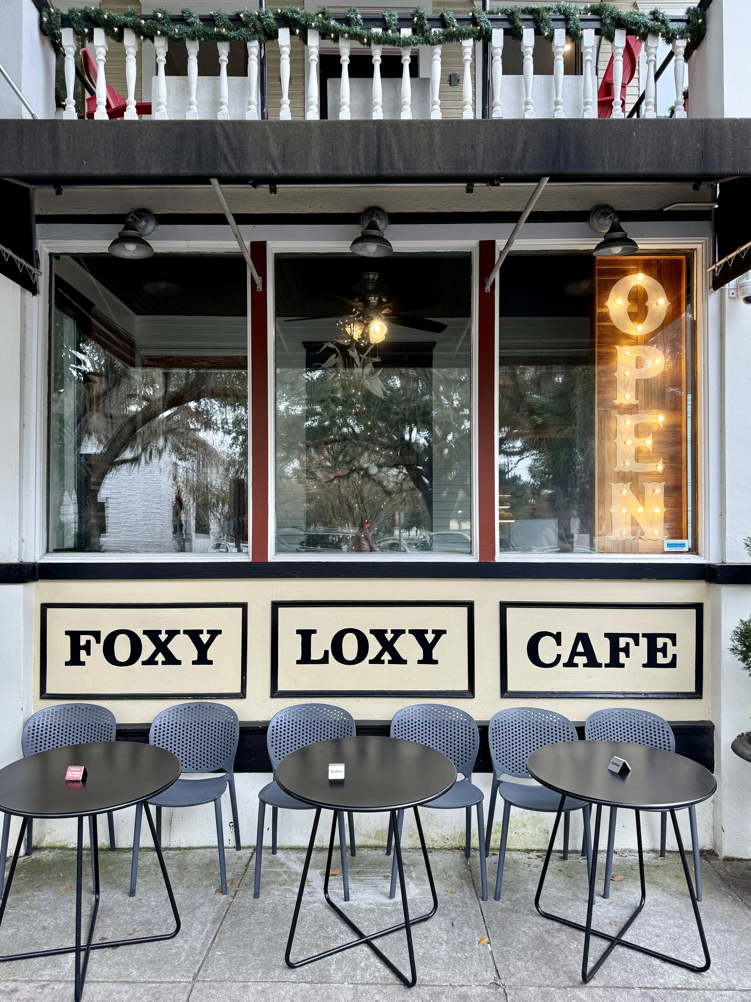 Front of Foxy Loxy Cafe, with a glowing Open sign, and small round black tables and chairs in Savannah, GA