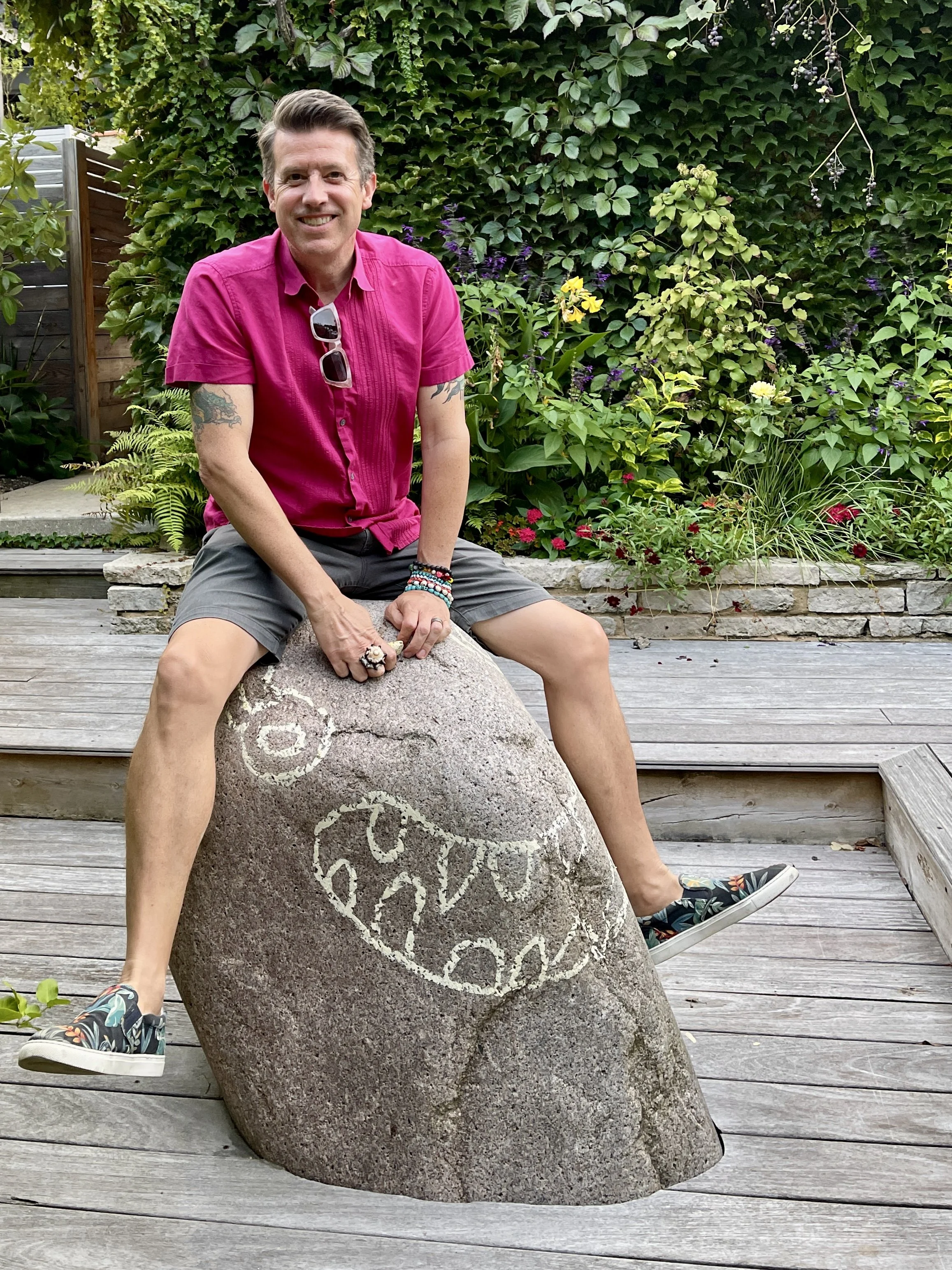 Man in pink shirt and gray shorts sits atop rock with smiling face drawn on it