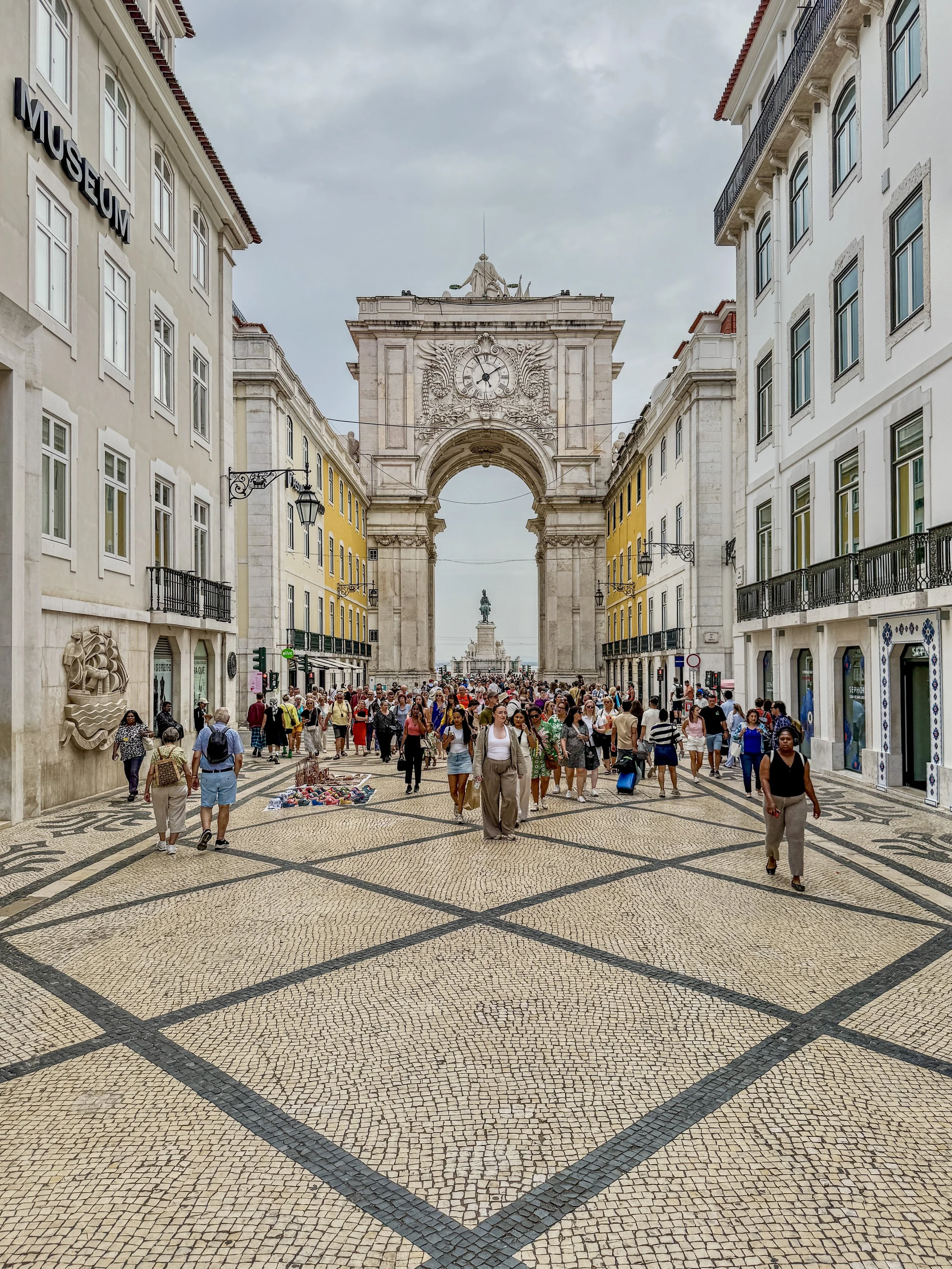 People crowd around the Rua Augusta Arch atop tiled pedestrian walkway in the Baixa neighborhood in Lisbon, Portugal
