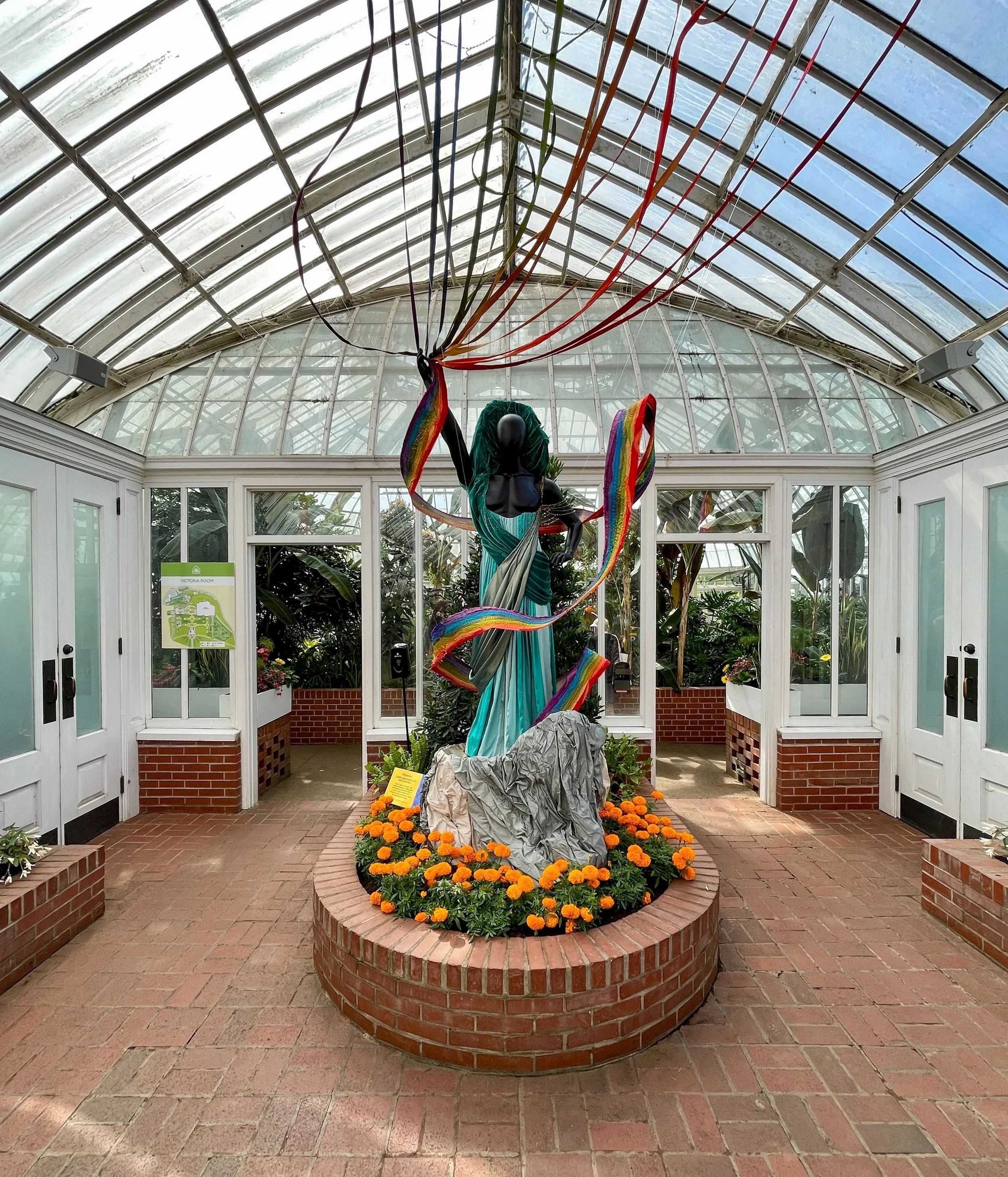 Mannequin wearing blue dress with rainbow fabric flowing around it in a planter filled with orange marigolds in glass conservatory at the Phipps