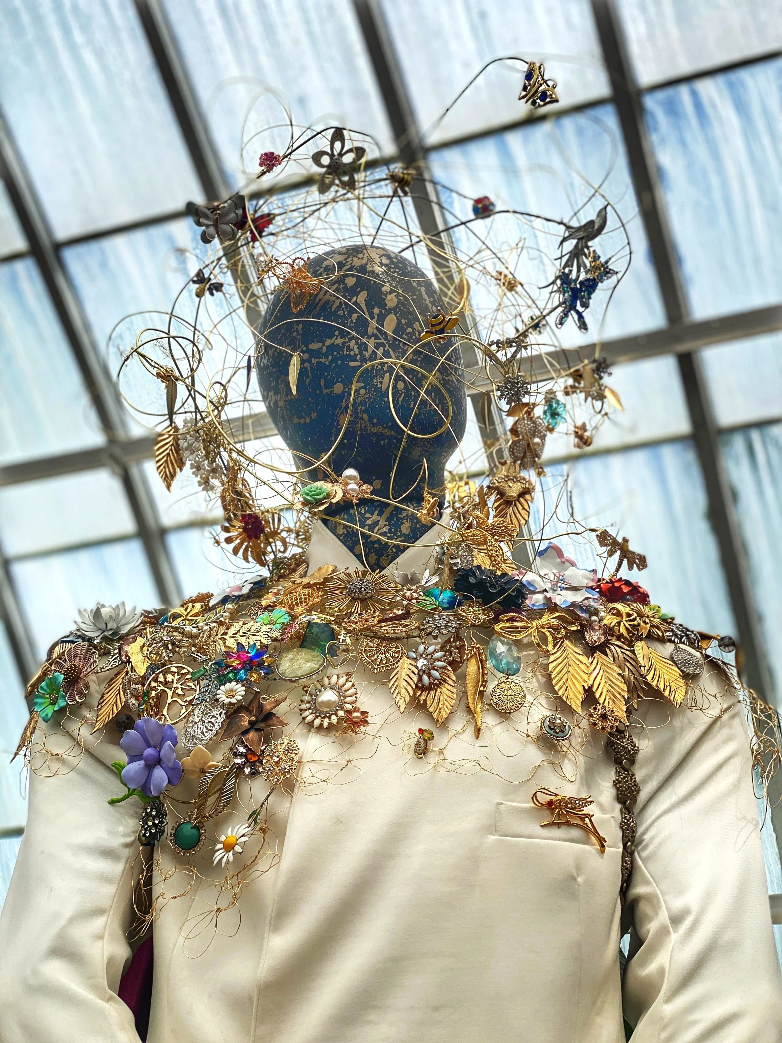 Closeup of mannequin head wearing twisted gold wire strung with metal leaves, flowers, brooches and other jewelery