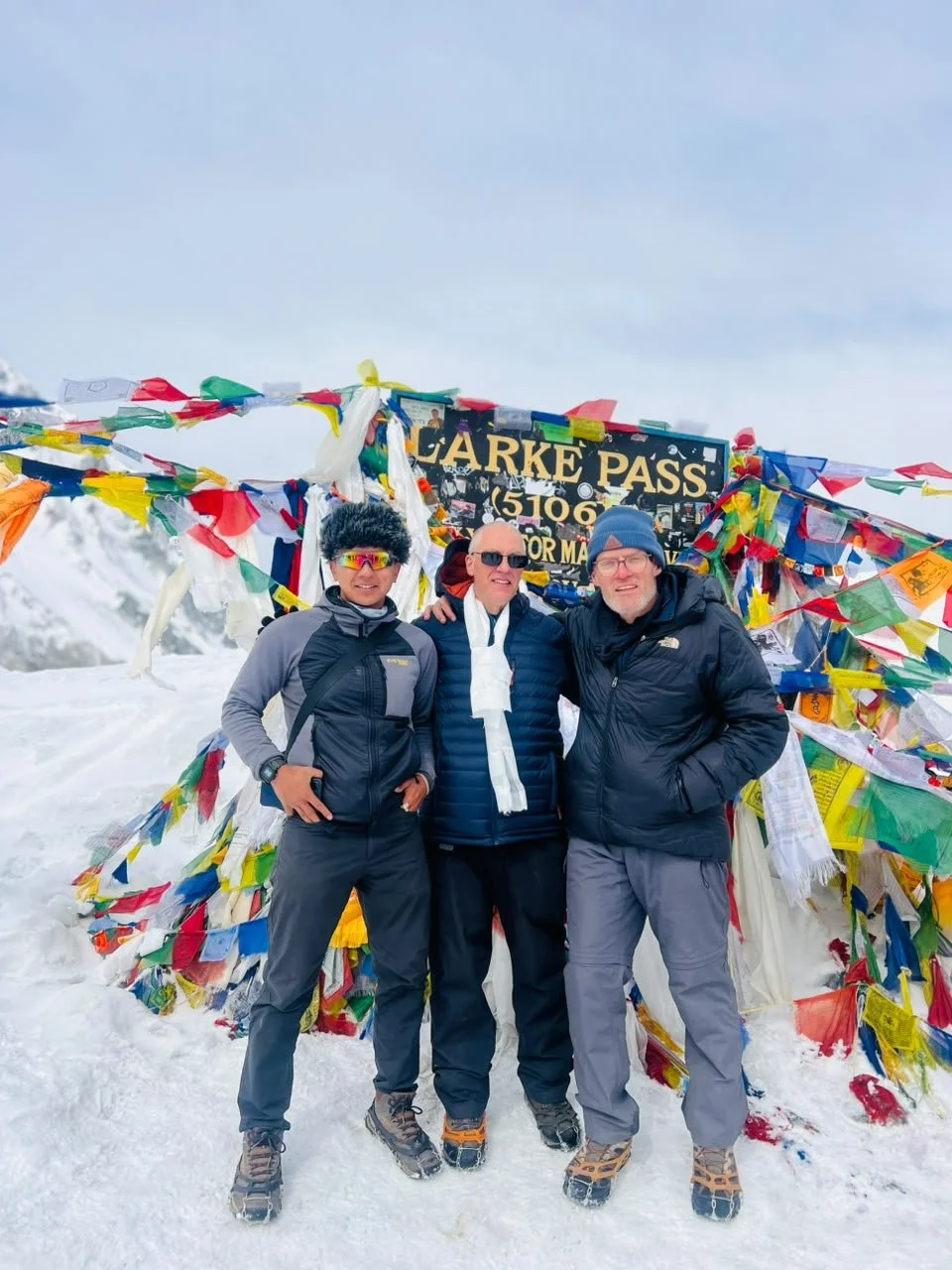 Three men stand in the snow by the colorful prayer flags at Larke La Pass in Nepal