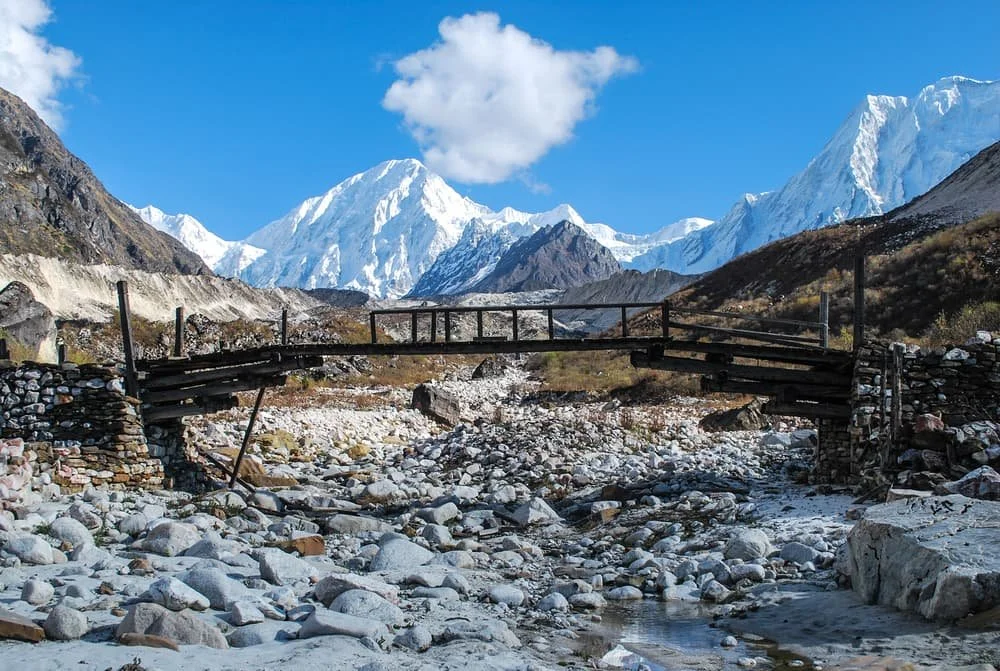 A wooden bridge covers snow-covered rocks in a river bed with mountains all around along the Manaslu Circuit trek in Nepal