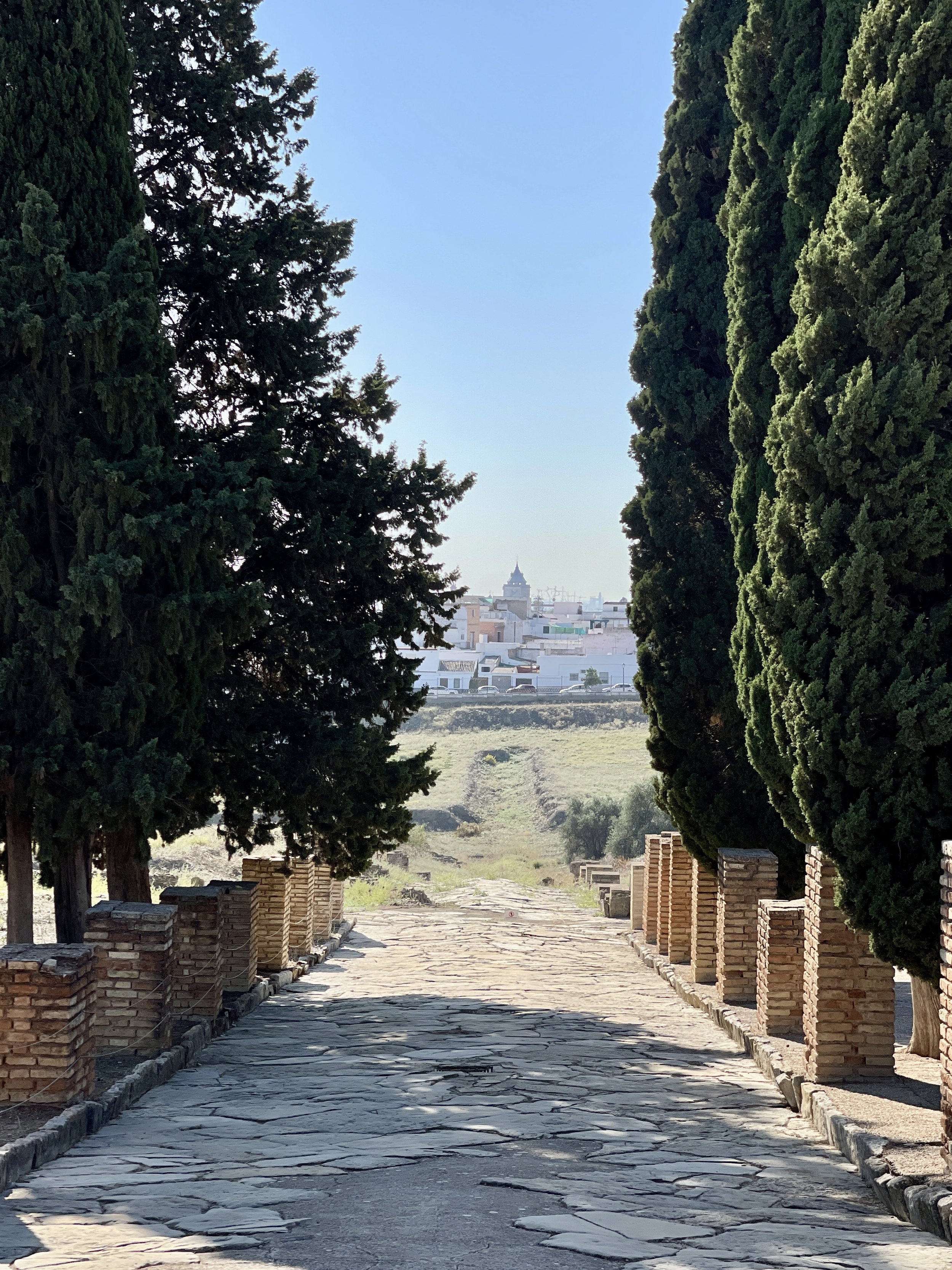A pathway lined with brick columns and cypresses leads to the town of Santiponce