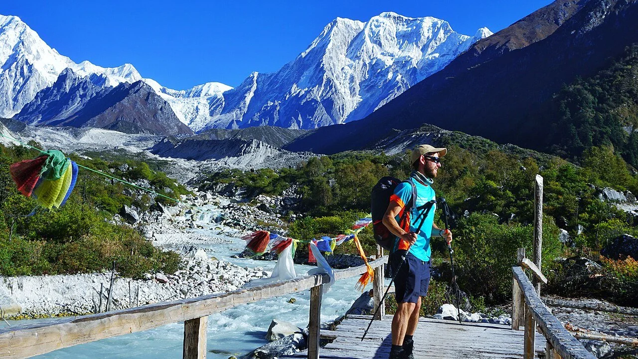A man hikes the Manaslu Circuit trek in Nepal on a wood bridge with snow-covered mountains in the background