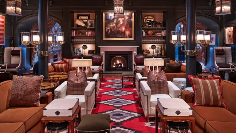Brown couches, white chairs, red carpet with gray diamonds and a fireplace in the lobby of Hotel Jerome in Aspen