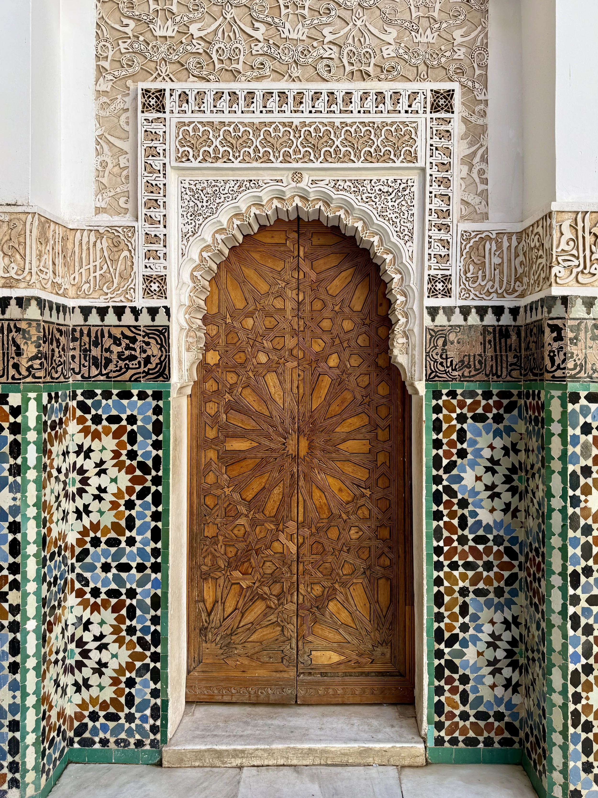 Carved Arabic calligraphy and patterns in plasterwork, a wooden door and zellij tiles at Ben Youssef Madrasa