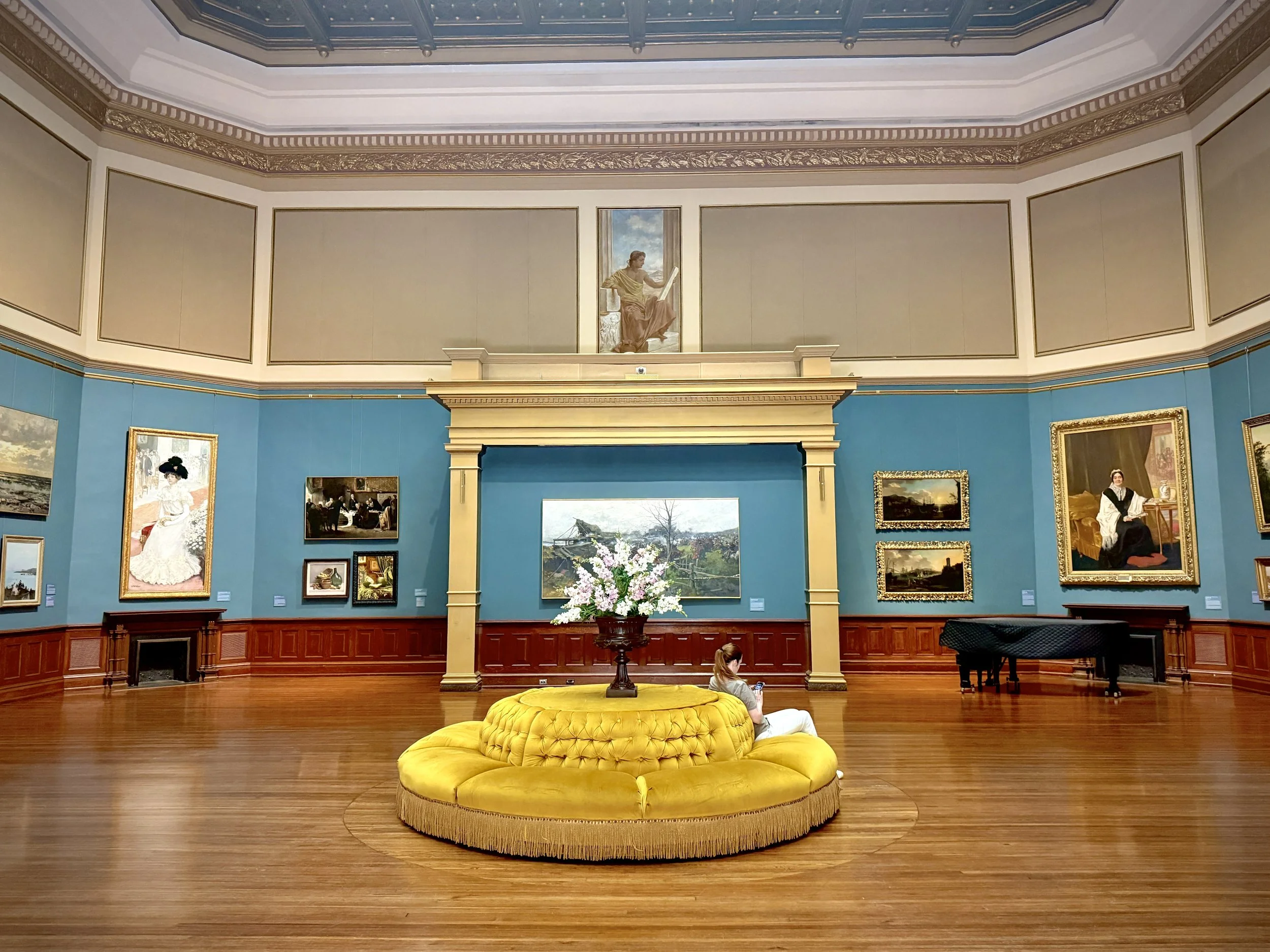 The Rotunda Gallery at the Telfair Academy in Savannah, Georgia, with blue walls covered with paintings, and a yellow circular settee with a vase of flowers atop it and a woman sitting on it, looking at her phone