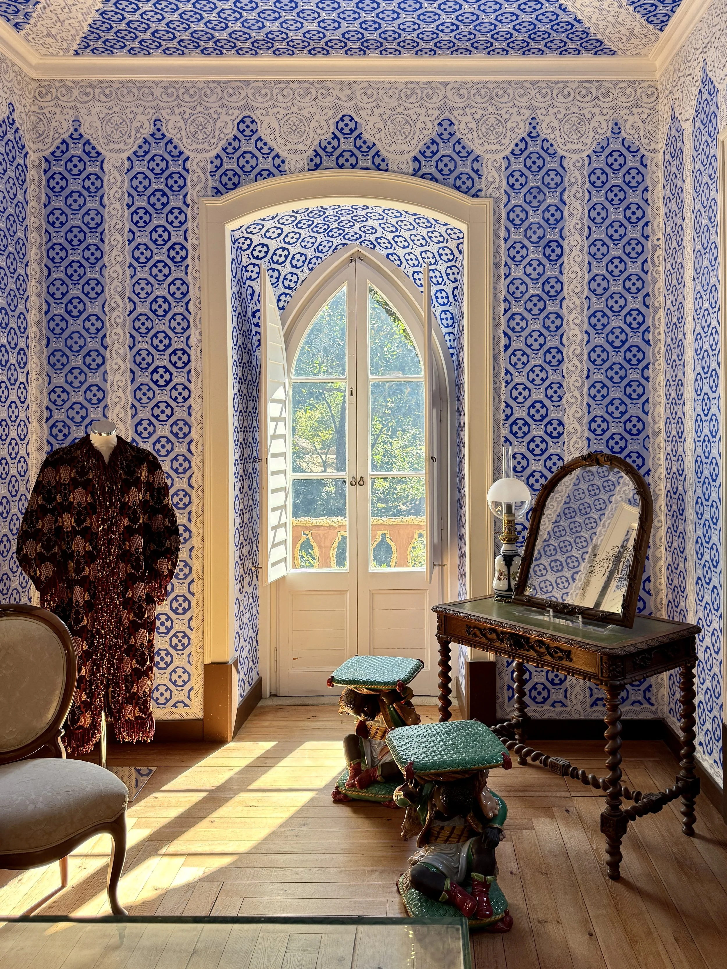 A blue room with dressing down, arched window, stools, dressing table with mirror, chair and blue patterned walls at the Chalet da Condessa d’Edla in Sintra
