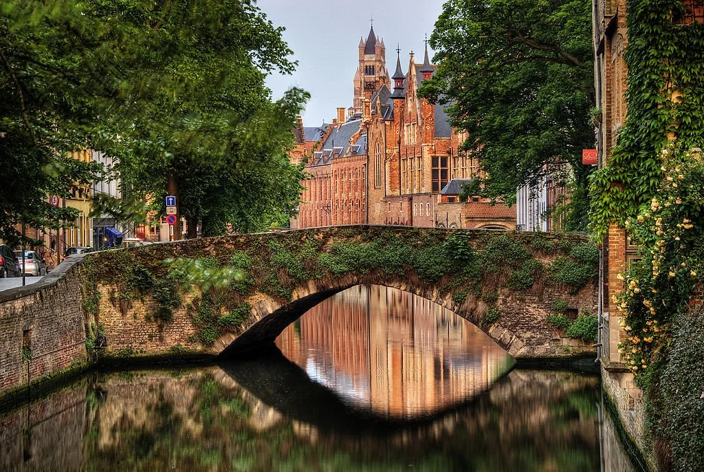 Rose-colored buildings along a tree-lined river with a cute plant-covered bridge in Bruges, Belgium