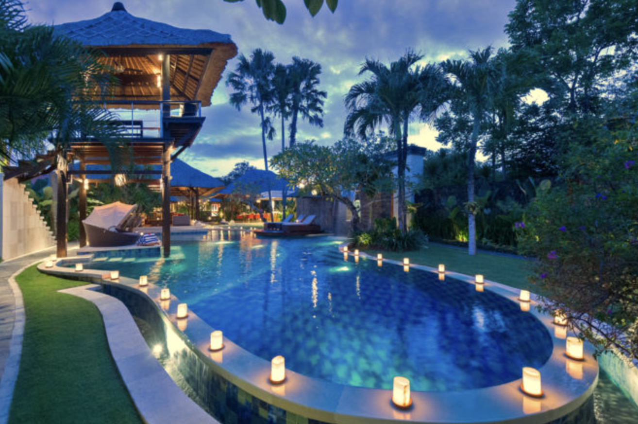A pool ringed by lights with palm trees and a wooden overlook structure at twilight on Bali