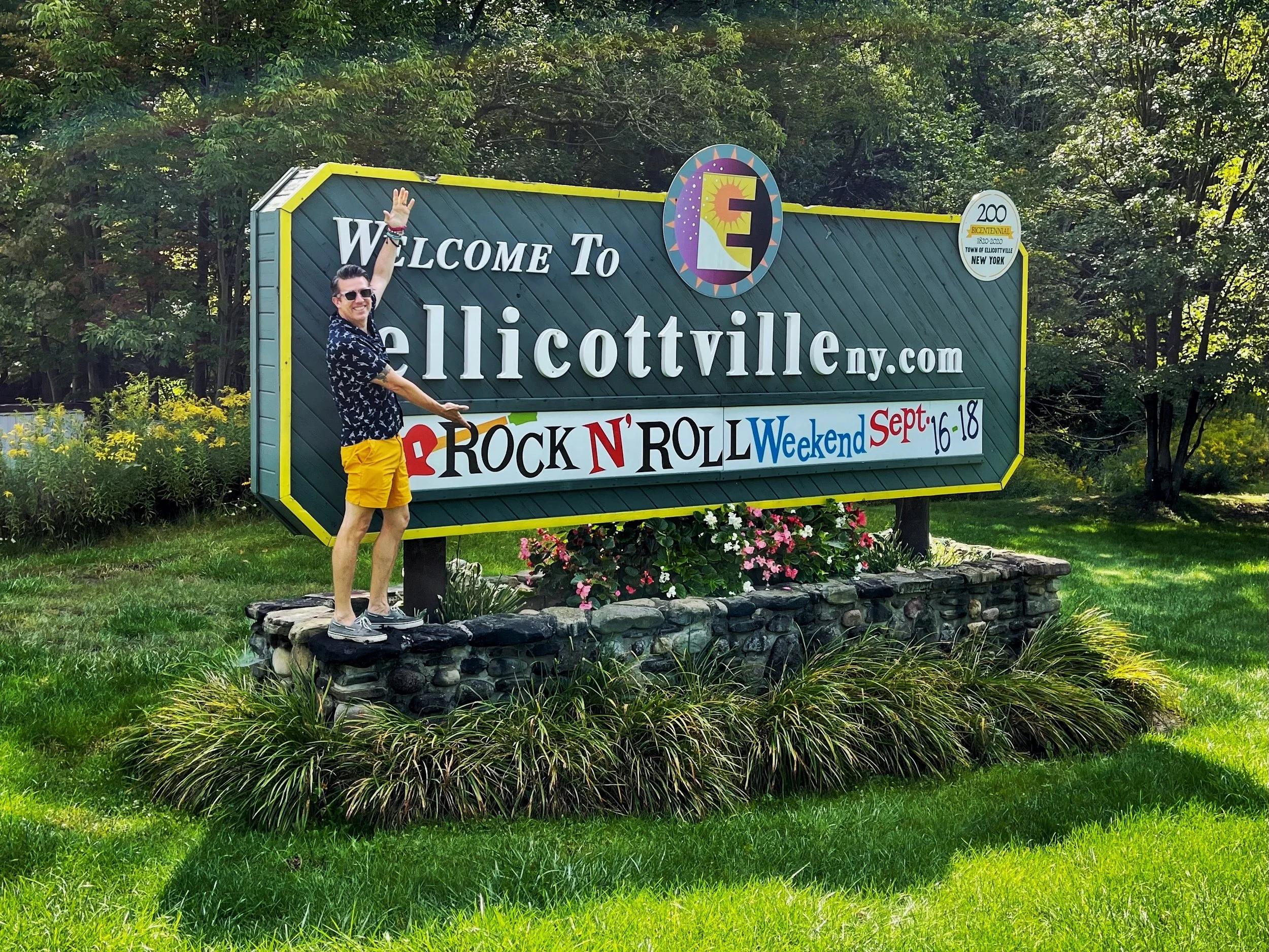 Man in yellow shorts by Welcome to Ellicottville sign