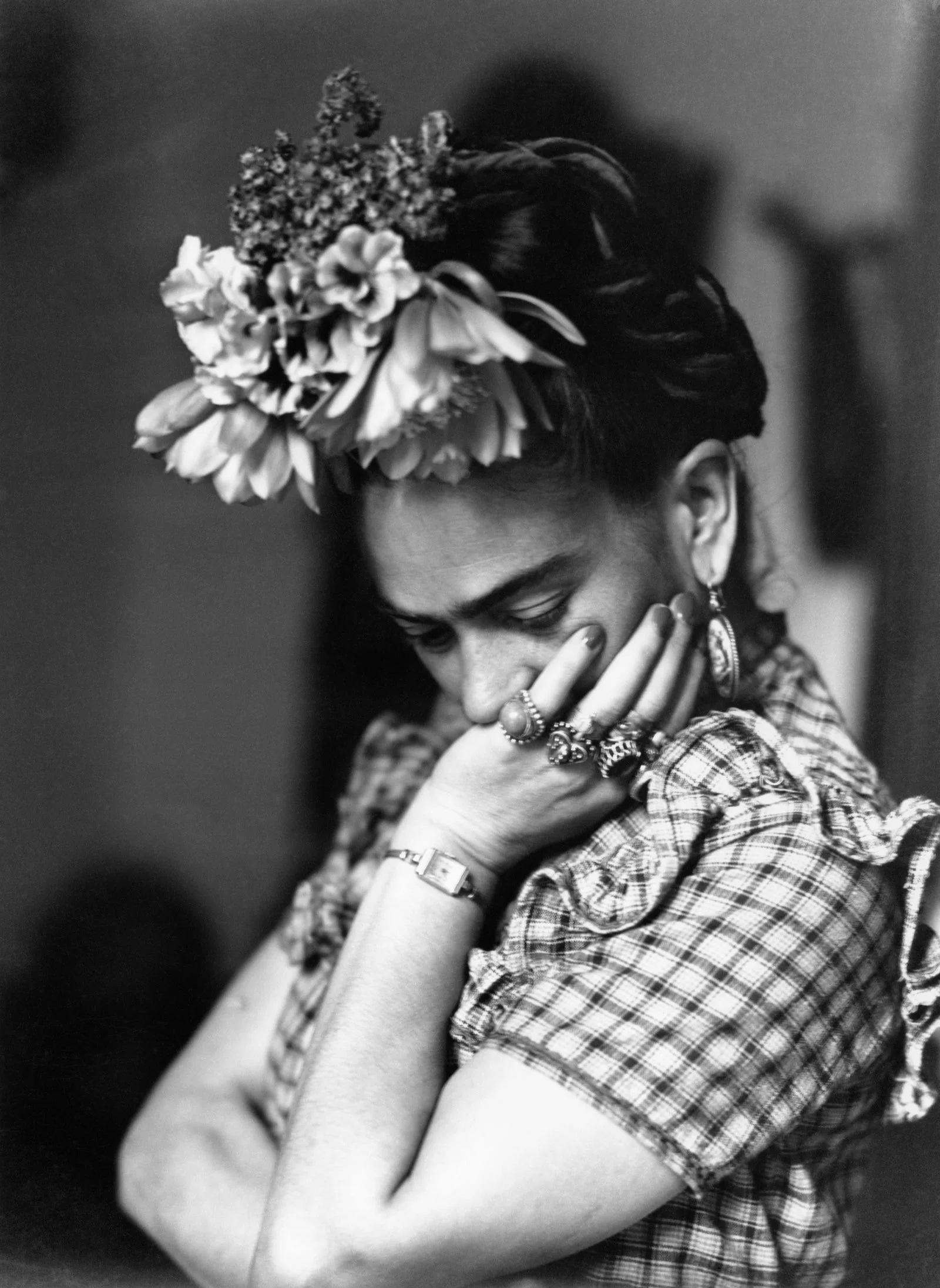 Frida Kahlo, wearing lots of rings and a floral headpiece, has her hand on her face and looks down in a sad manner