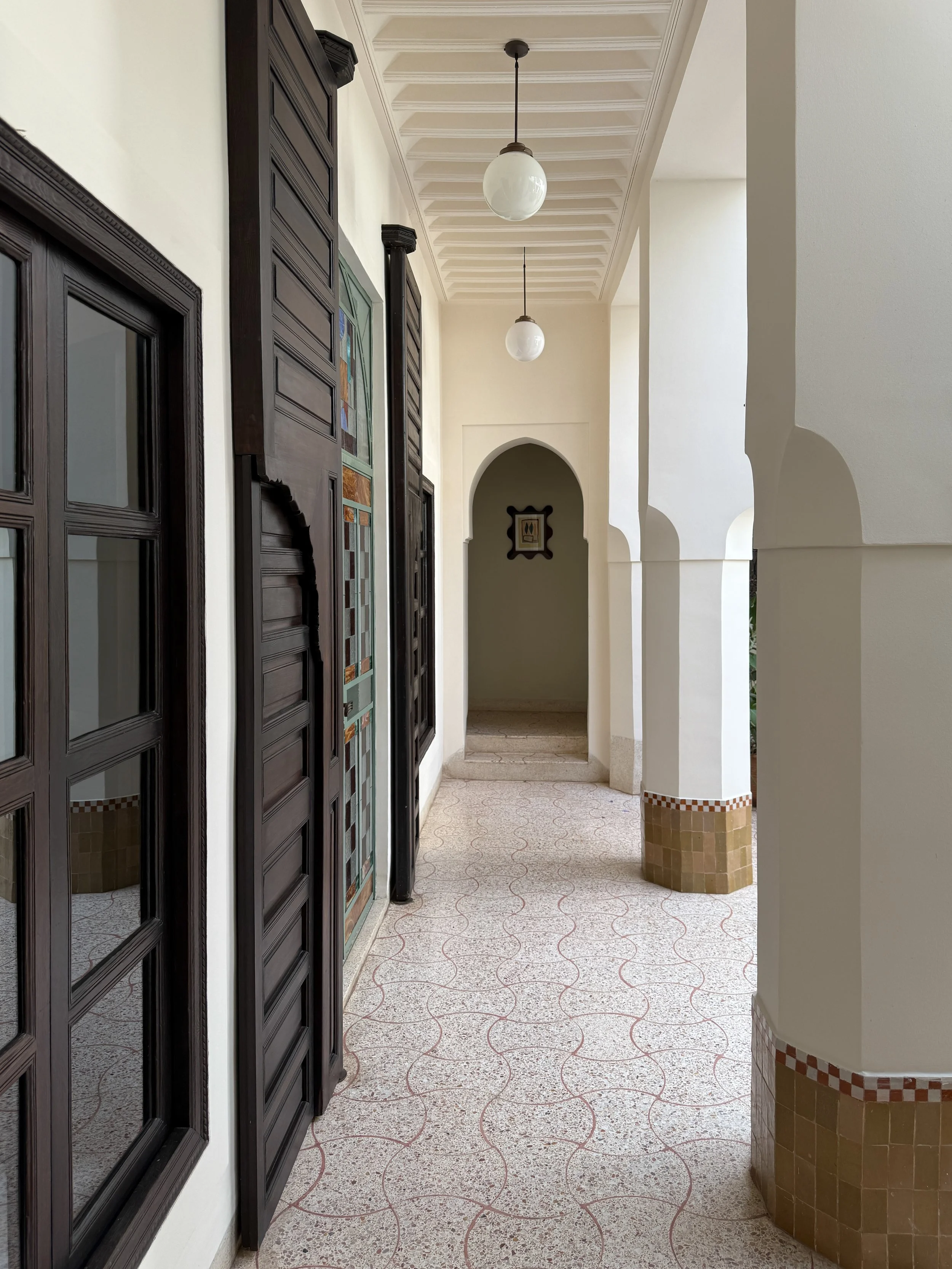A hallway with white columns and dark wood doors and window frames at Rosemary Riad in Marrakech, Morocco