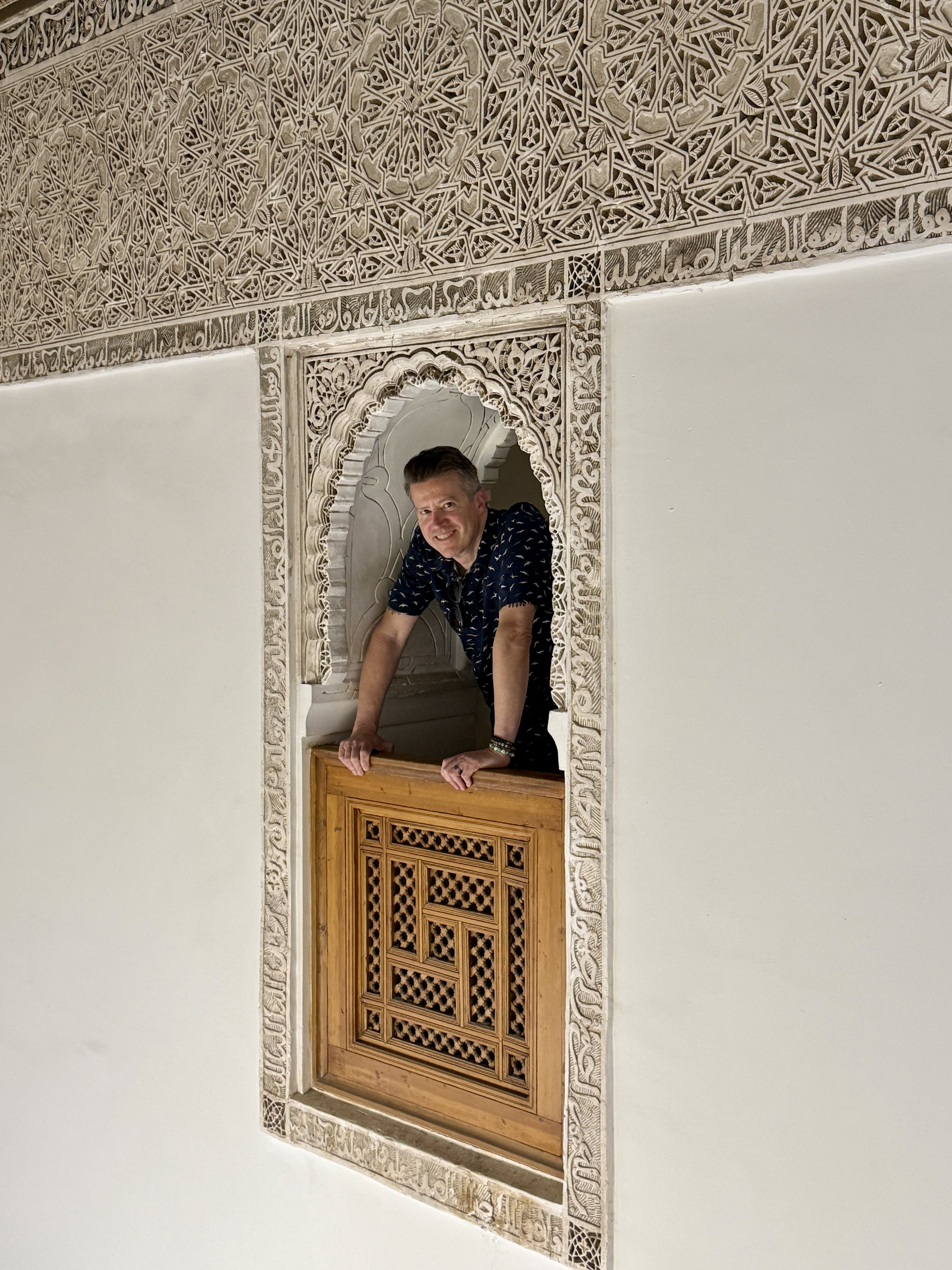 A man leans on a wood balcony in a carved arched interior window upstairs at Ben Youssef Madrasa