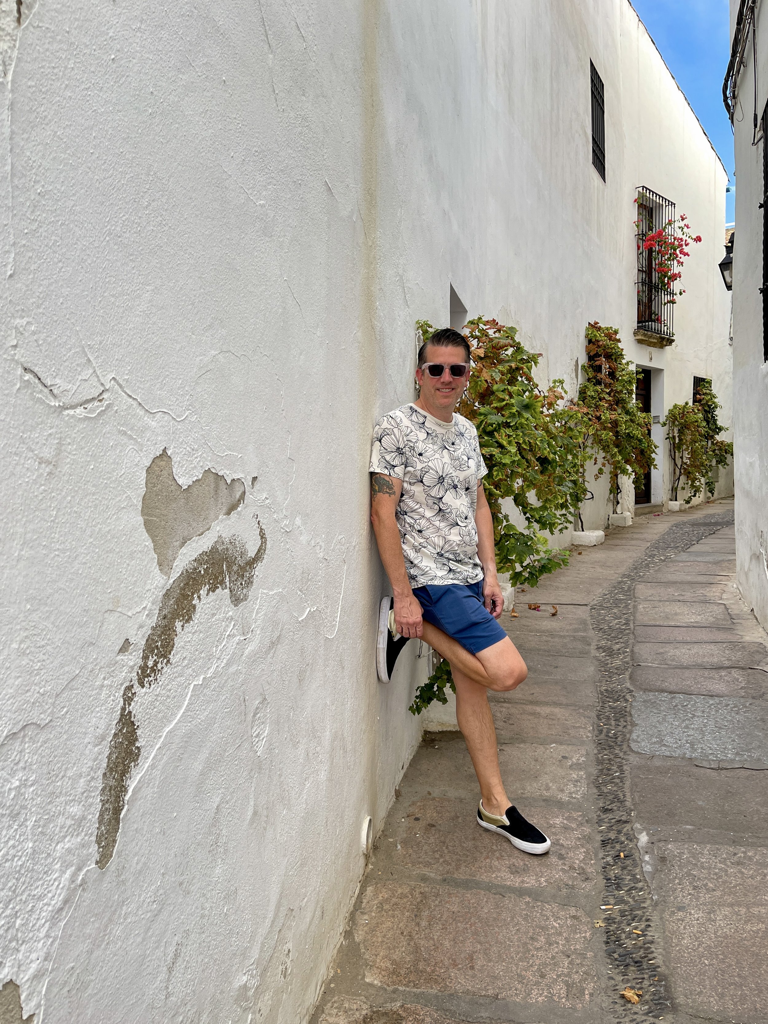 Man in floral patterned t-shirt and blue shorts leans against white wall with greenery on narrow lane in Cordoba, Spain