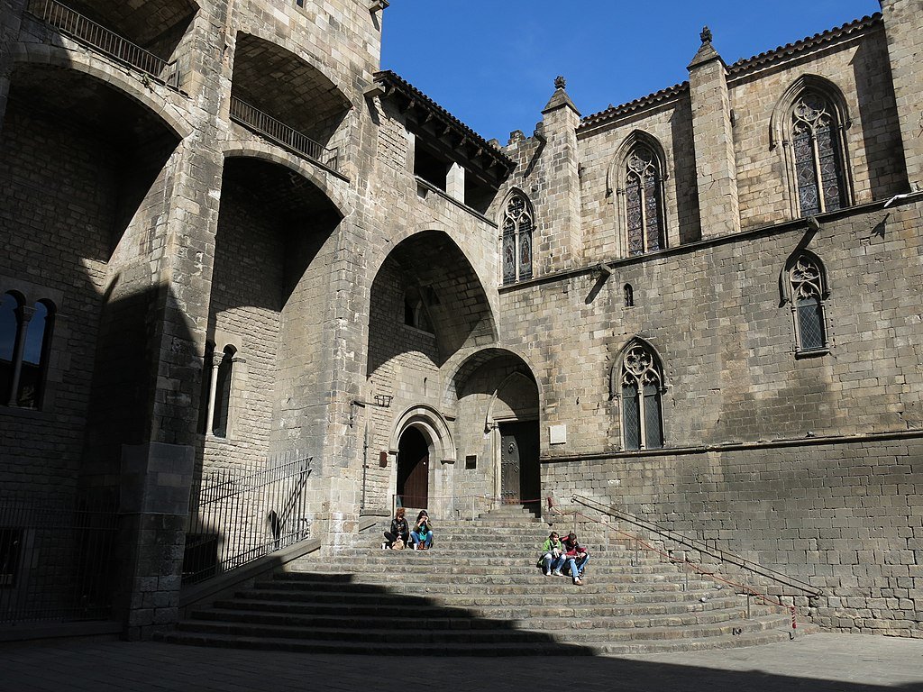People sit on the steps of the gray stone building in Plaça del Reí  in Barcelona