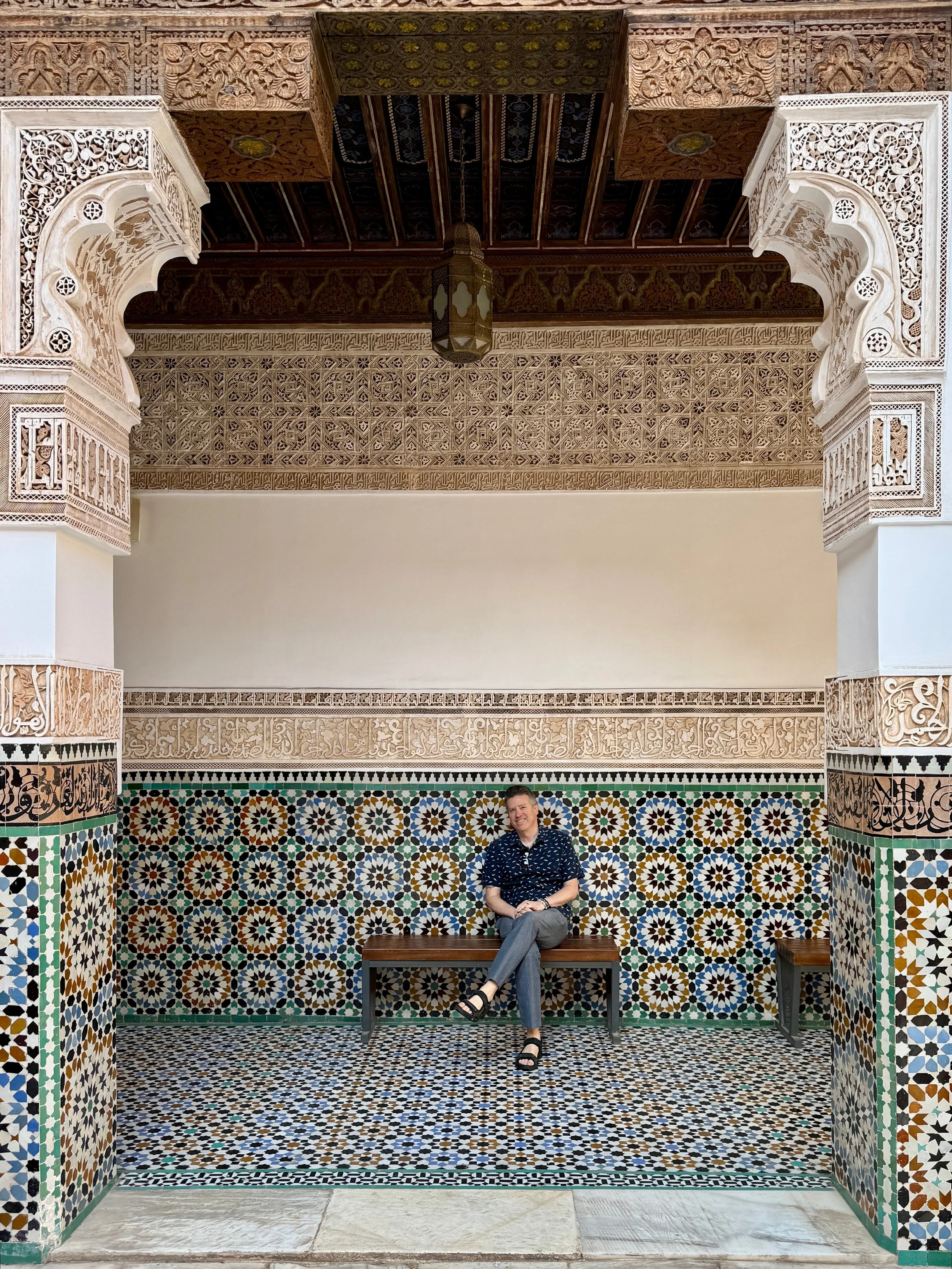 A man sits on a bench among colorful zellij tiles at Ben Youssef Madrasa,