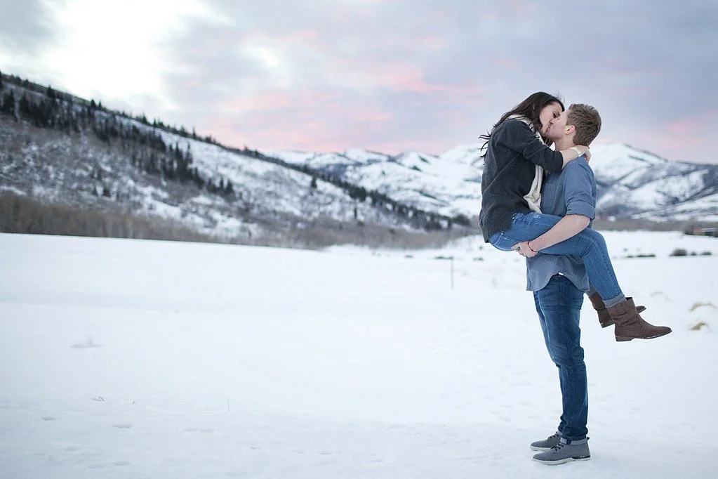A man holds up a woman by her legs as she wraps her arms around his head and kisses him by a snowy Aspen mountain