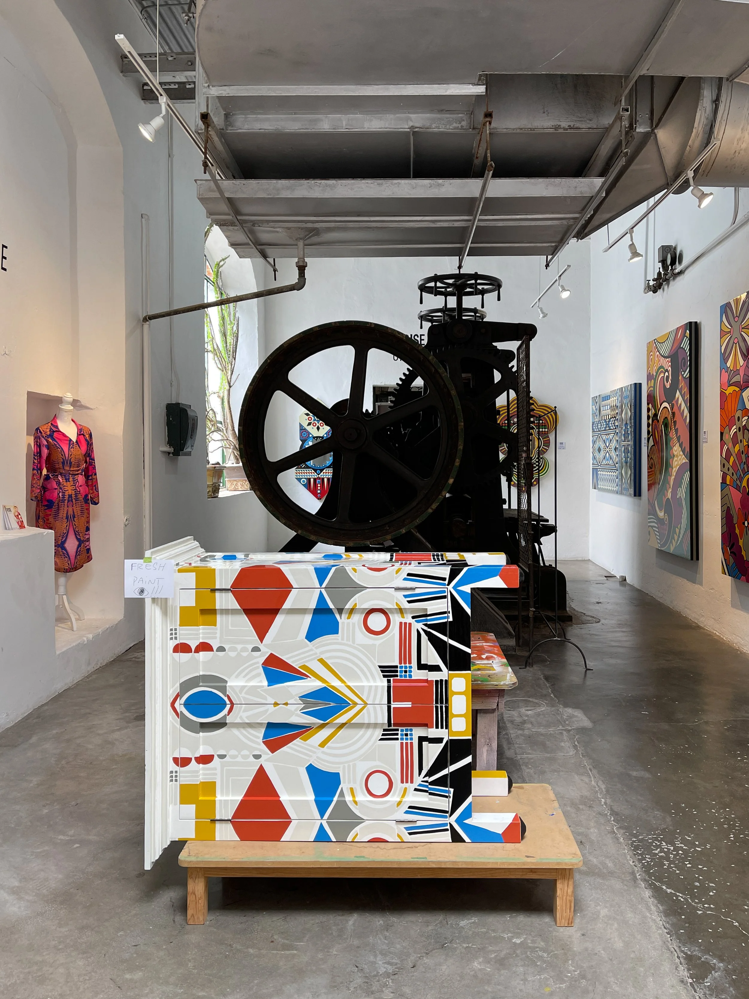 Rise gallery in Fabrica la Aurora with large piece of machine from the old textile factory, paintings, a pink dress and a wooden sculpture with primary colors in geometric designs