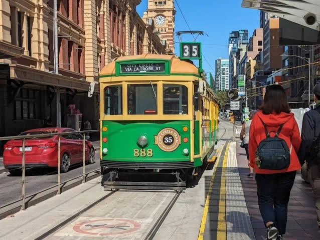 A woman walks along the sidewalk in Melbourne, Australia, as a green historic tram approaches