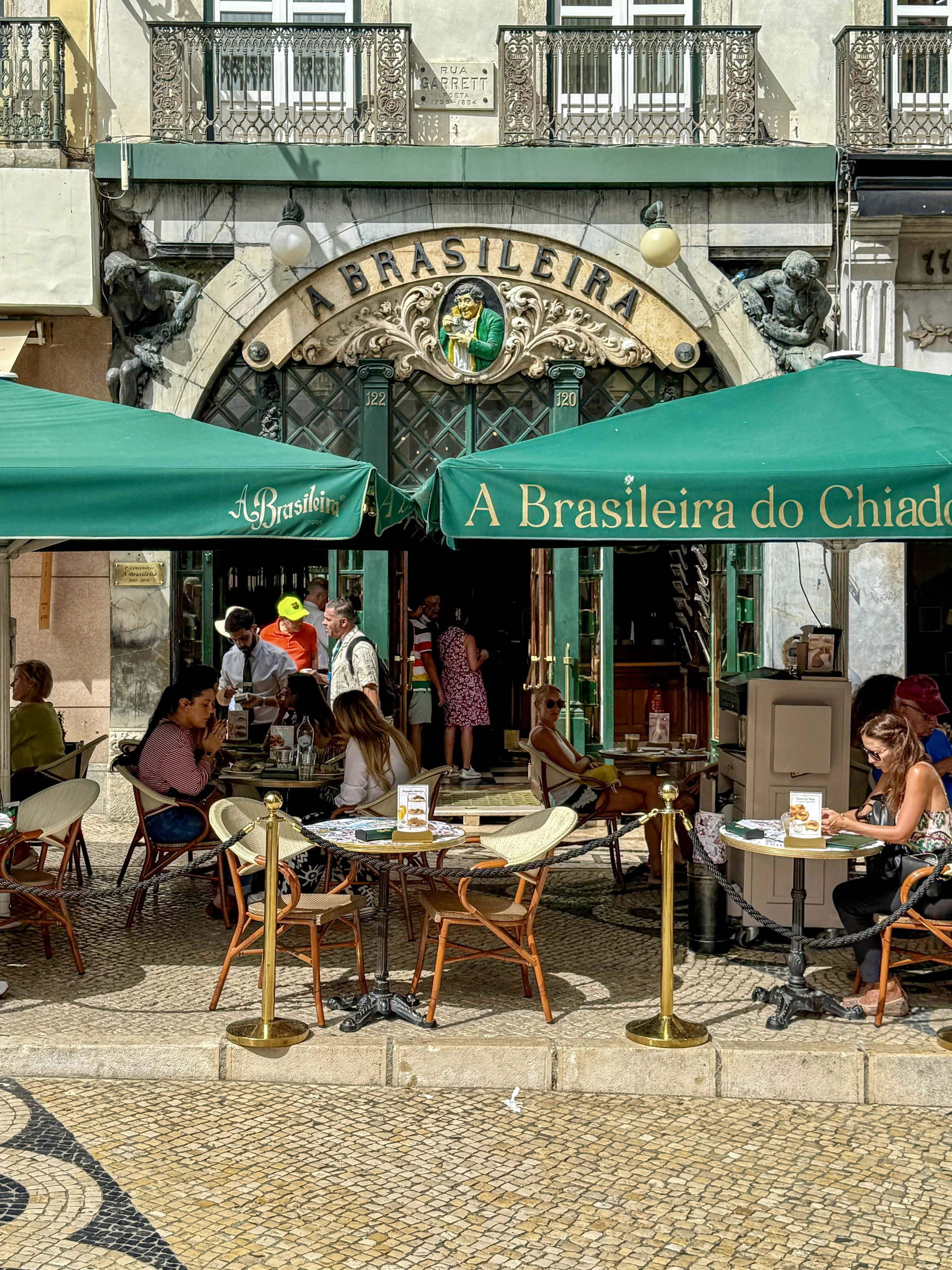 People sit at tables under green umbrellas at A Brasileira coffeeshop in Lisbon's Chiado neighborhood