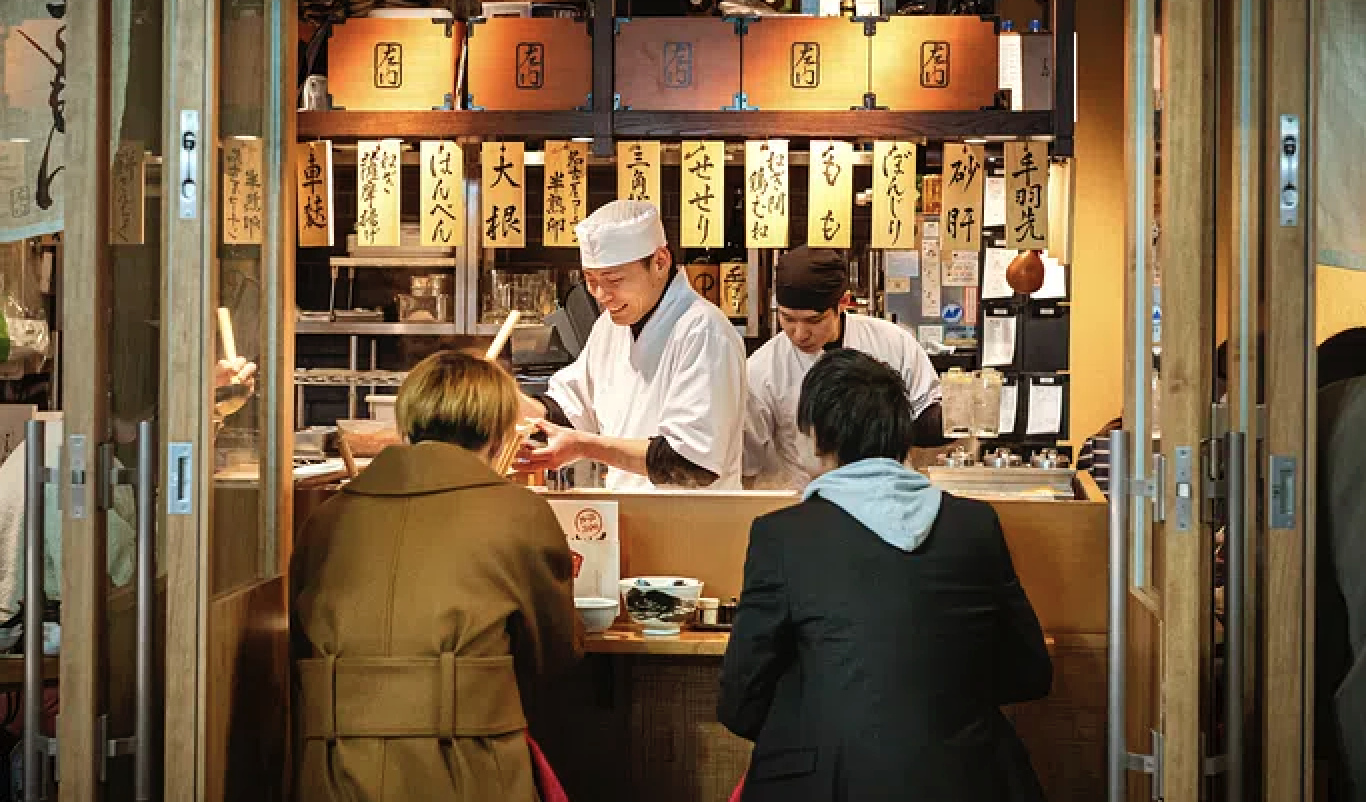 Chef smiles and serves food to woman and man at an izakaya pub