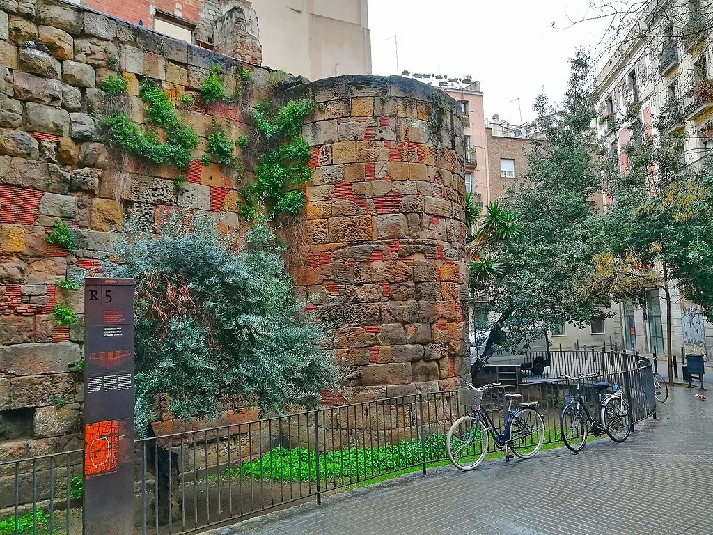 Two bikes parked along a fence by a reddish stone fortress wall in Plaça dels Traginers in Barcelona