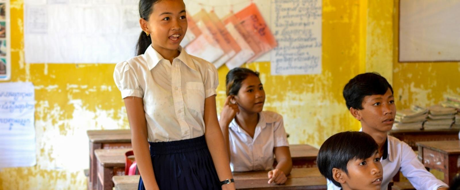 A girl in a white blouse stands up in classroom with faded yellow paint on the walls in Cambodia