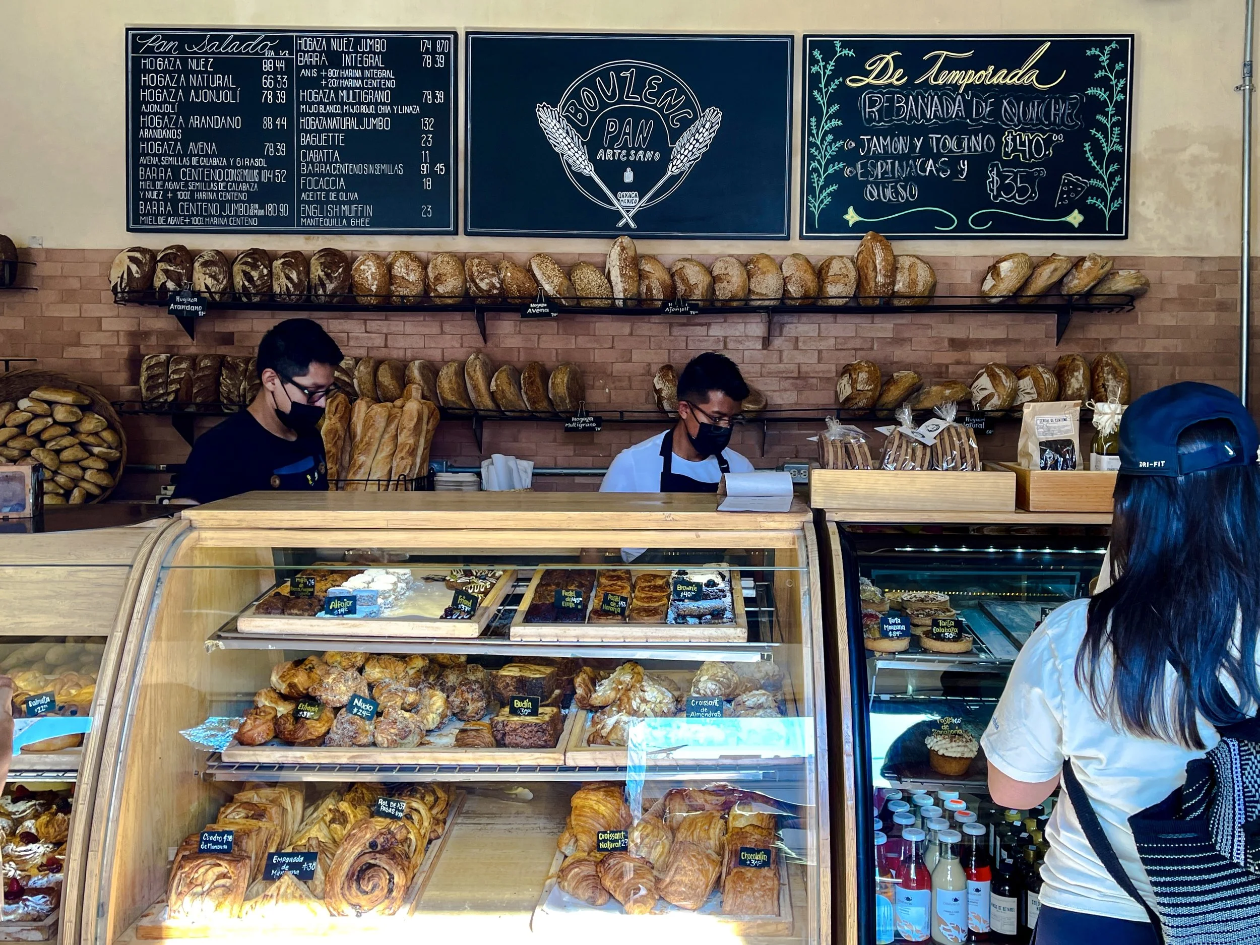 Two men in face masks behind the counter with display cases filled with pastries and a wall with blackboard menus and shelves with bread loaves, with dark-haired woman customer seen from behind