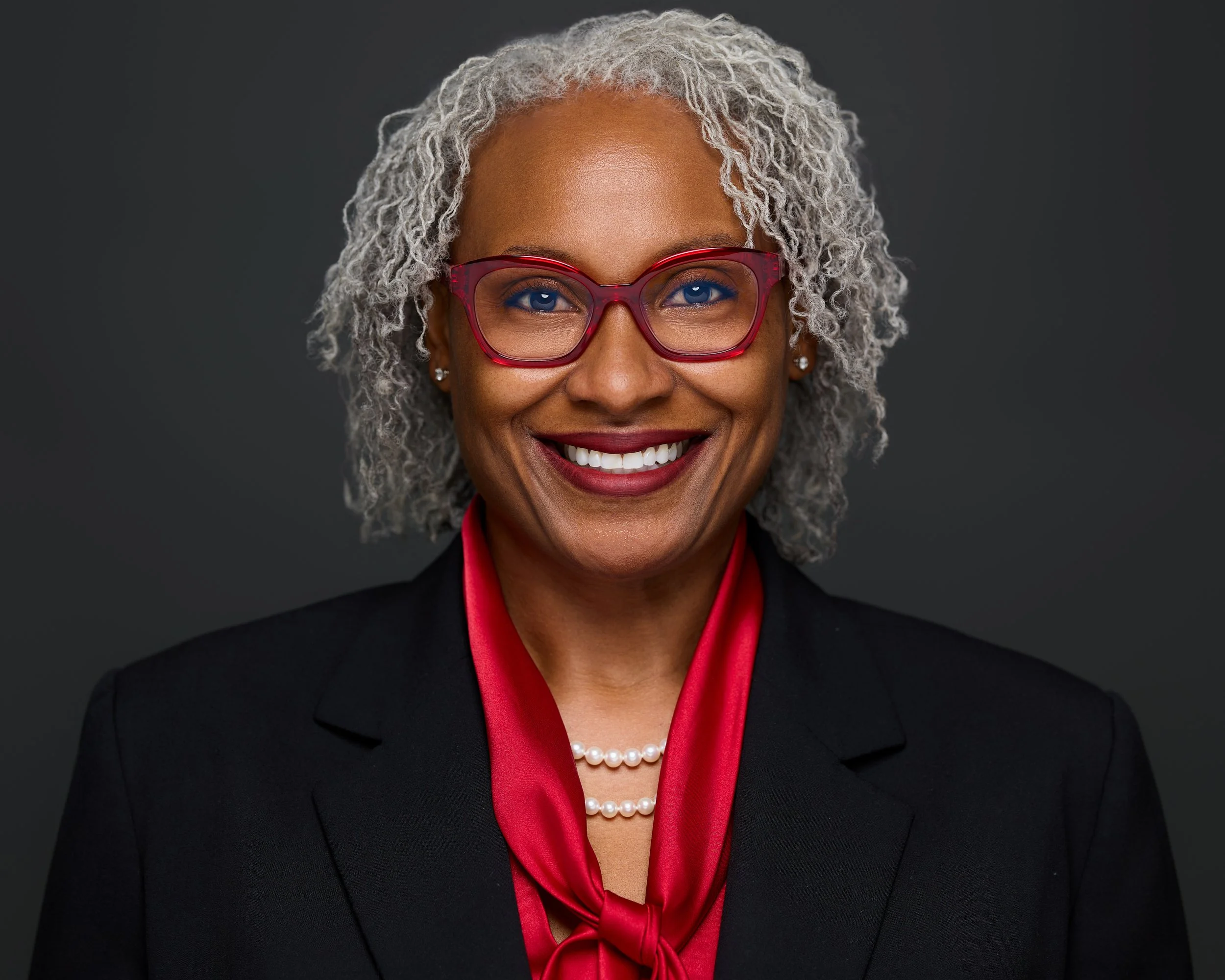 A smiling African American woman with gray curly hair, wearing red glasses, pearl earrings, a black blazer, and a red silk scarf, against a dark gray background.