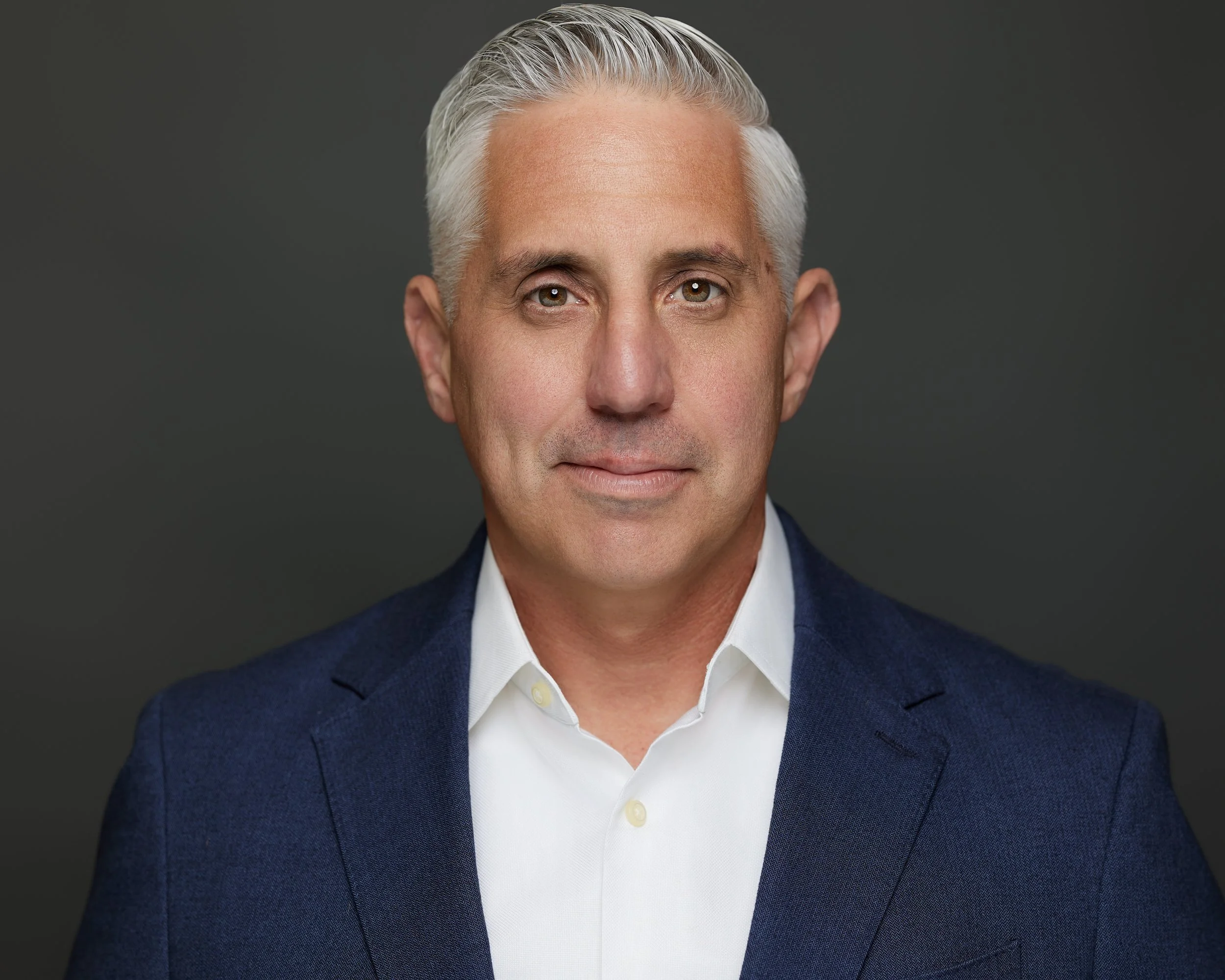 A professional headshot of a middle-aged man with silver hair, wearing a navy blue suit and white shirt, against a dark gray background.
