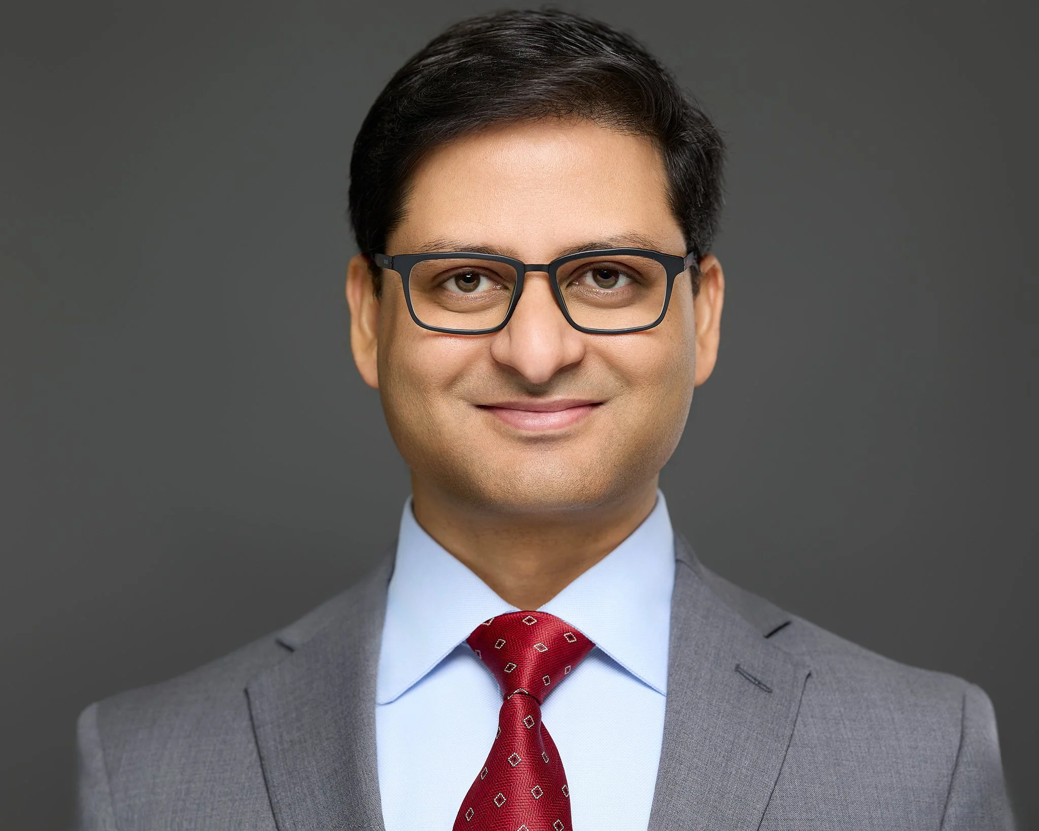 Professional man in gray suit, light blue shirt, red tie with small pattern, glasses, smiling against a dark gray background.