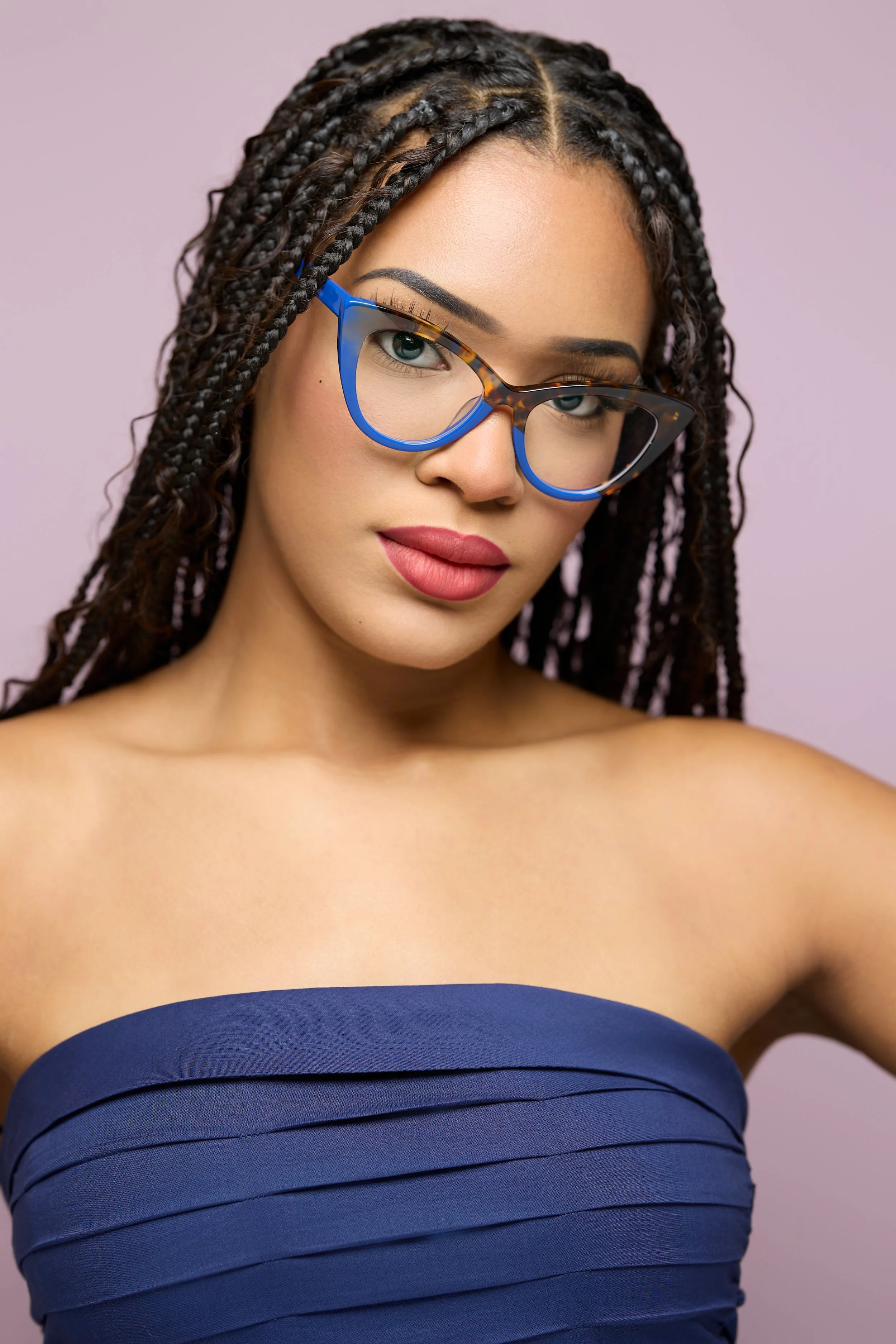 A woman with braided hair and glasses, wearing a strapless navy blue top, posing against a light purple background.