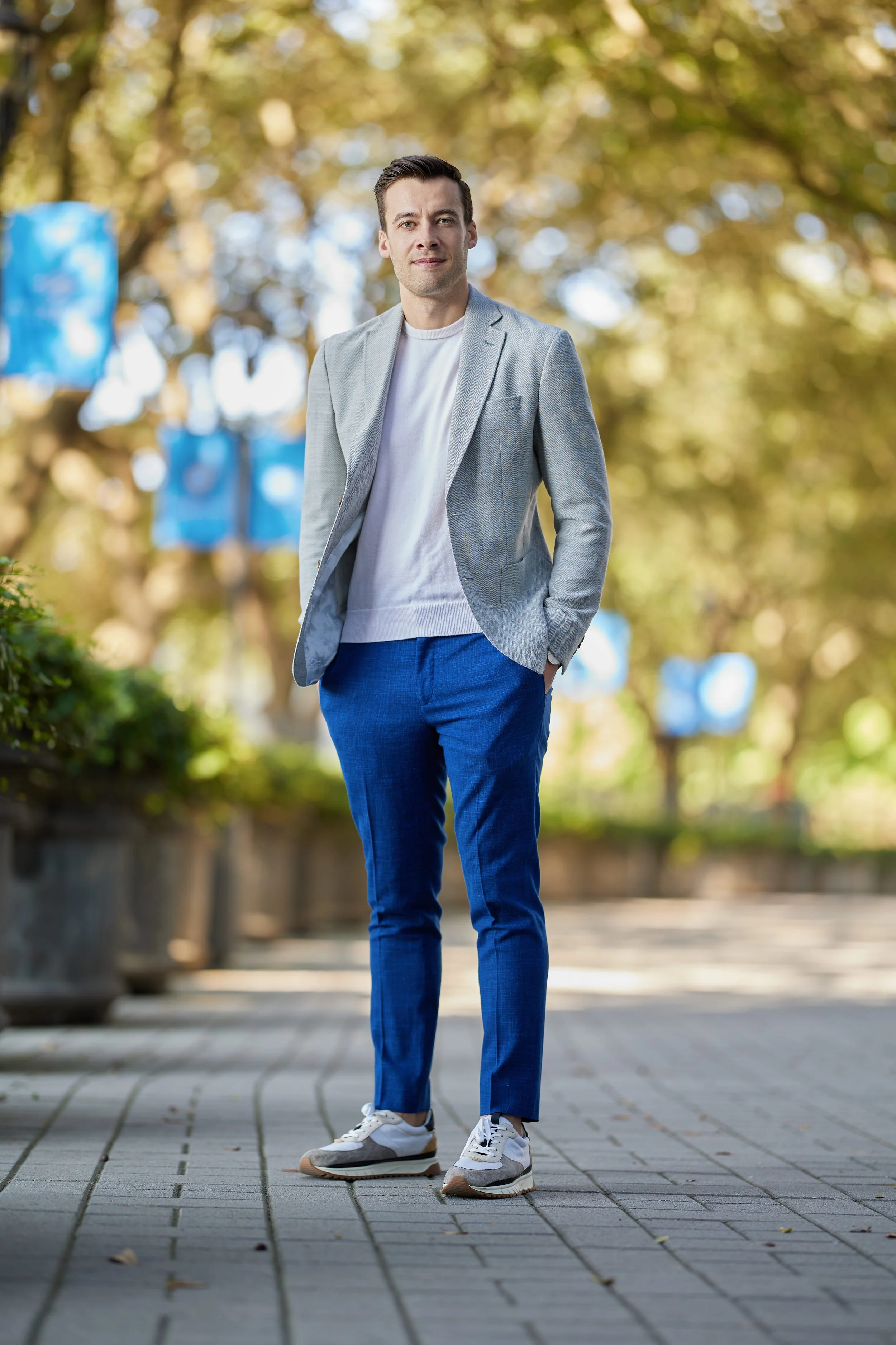 A man in a light gray blazer, white sweater, blue pants, and sneakers standing on a paved path outdoors with trees and blue flags in the background.
