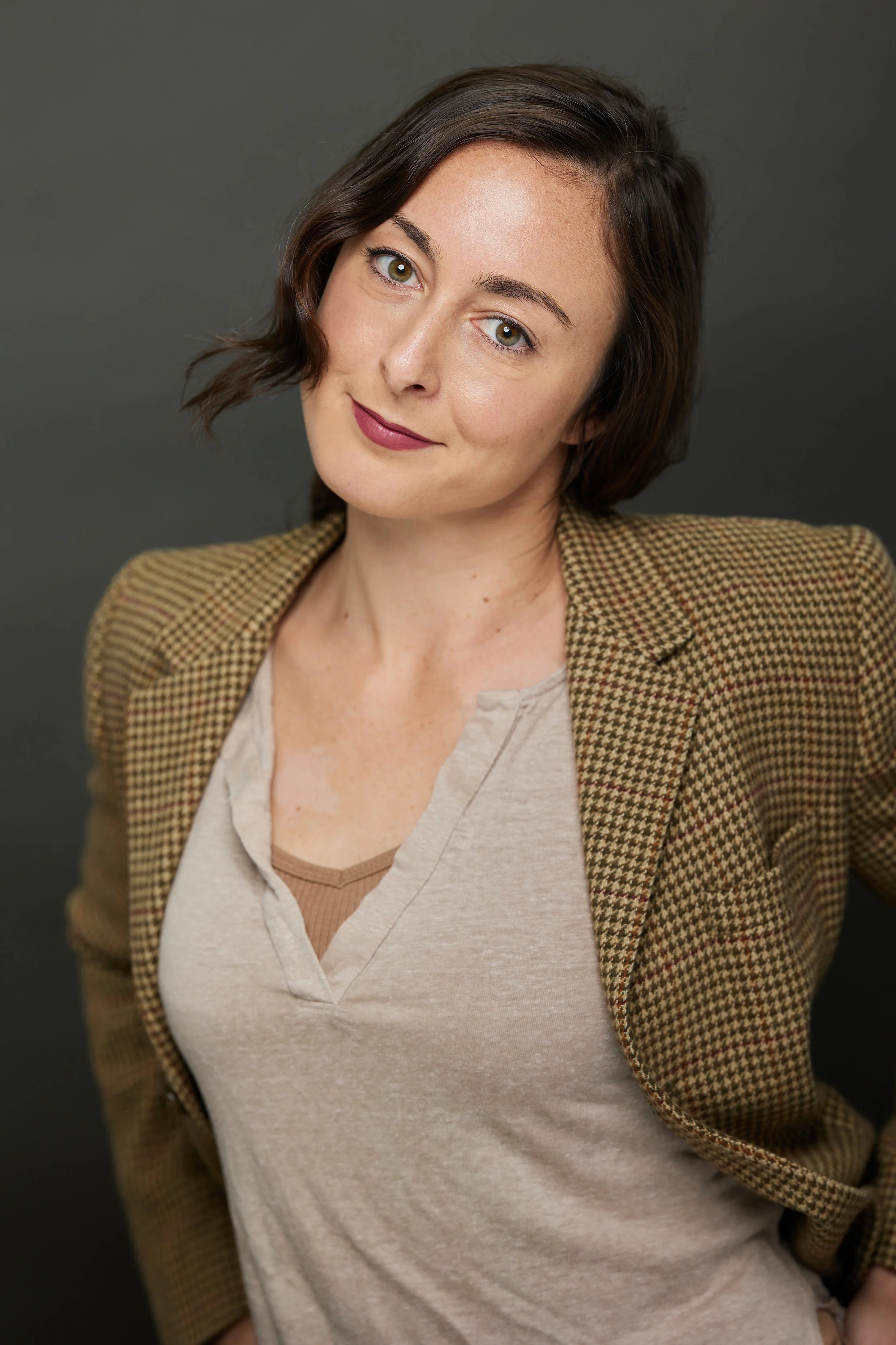 A woman with short brown hair, green eyes, and makeup, wearing a beige V-neck top and a brown checkered blazer, posing against a gray background.