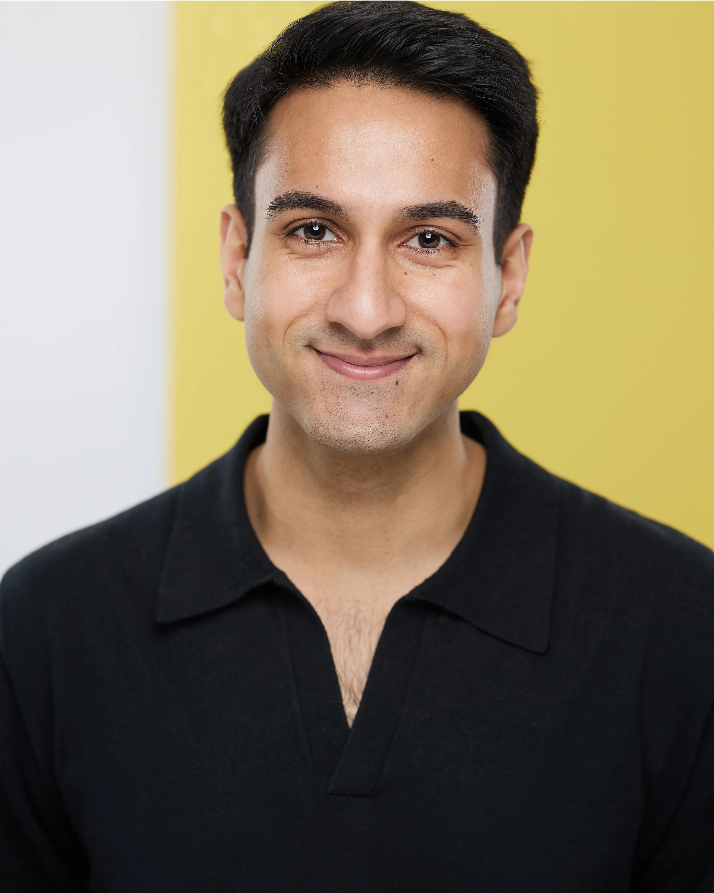 Close-up portrait of a young man with black hair, smiling, wearing a black shirt, with a yellow and white background.