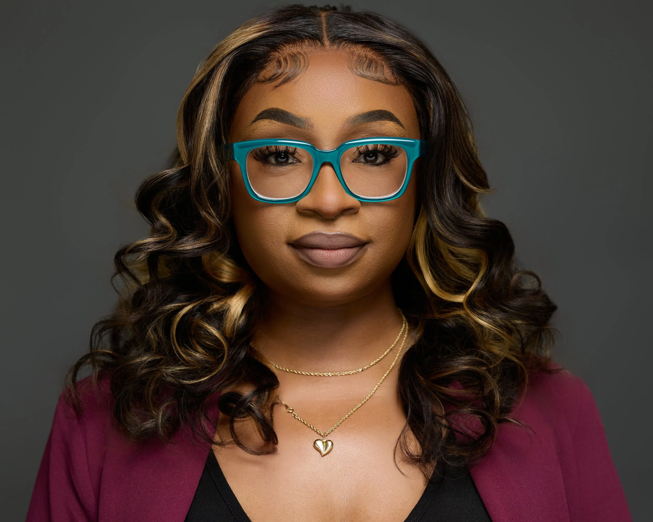 A woman with curly, highlighted hair wearing blue glasses, a maroon blazer, and gold jewelry, posing against a dark gray background.