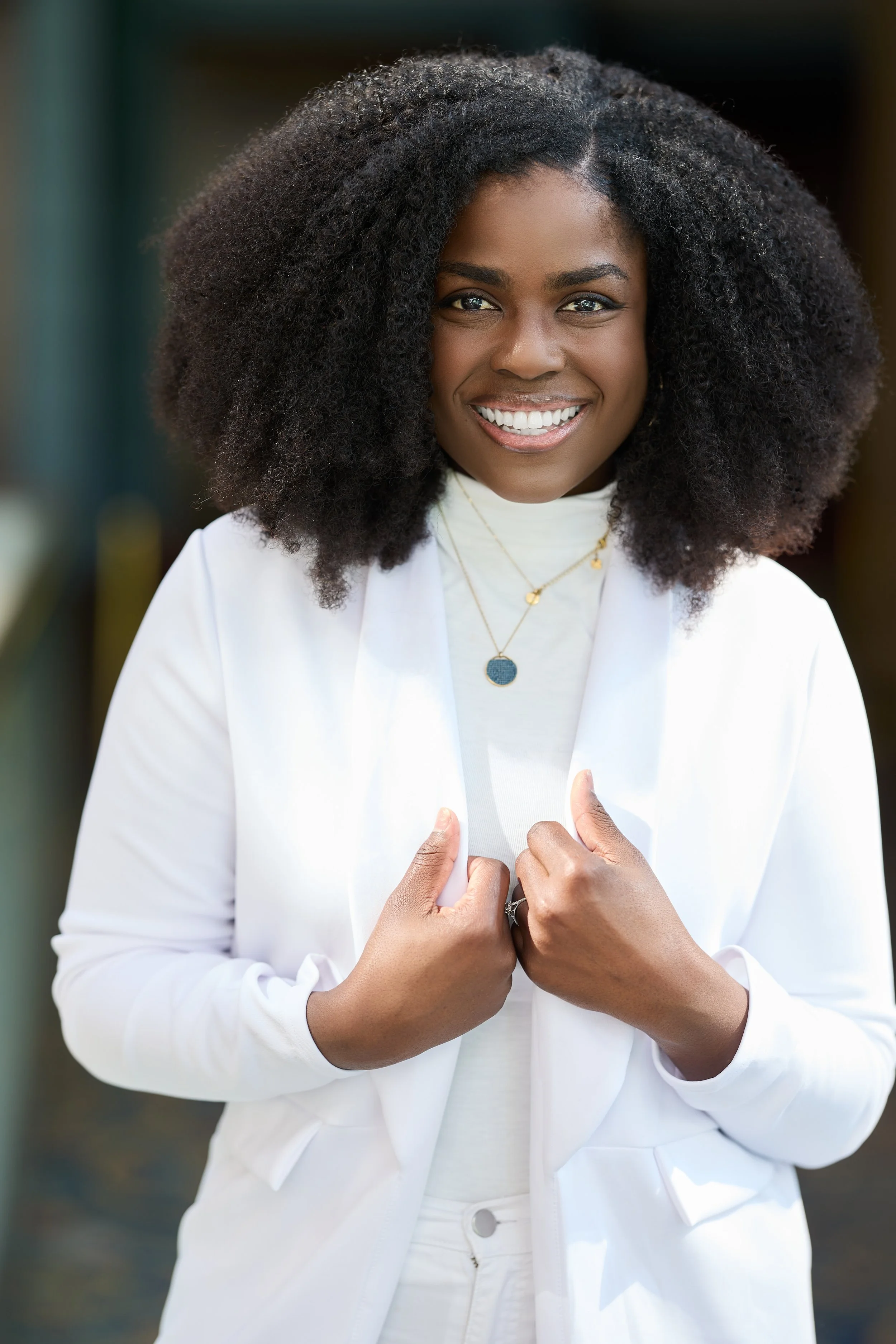 A woman with dark skin and curly black hair smiling and holding her white blazer with both hands, wearing a white top and gold jewelry, standing outdoors.