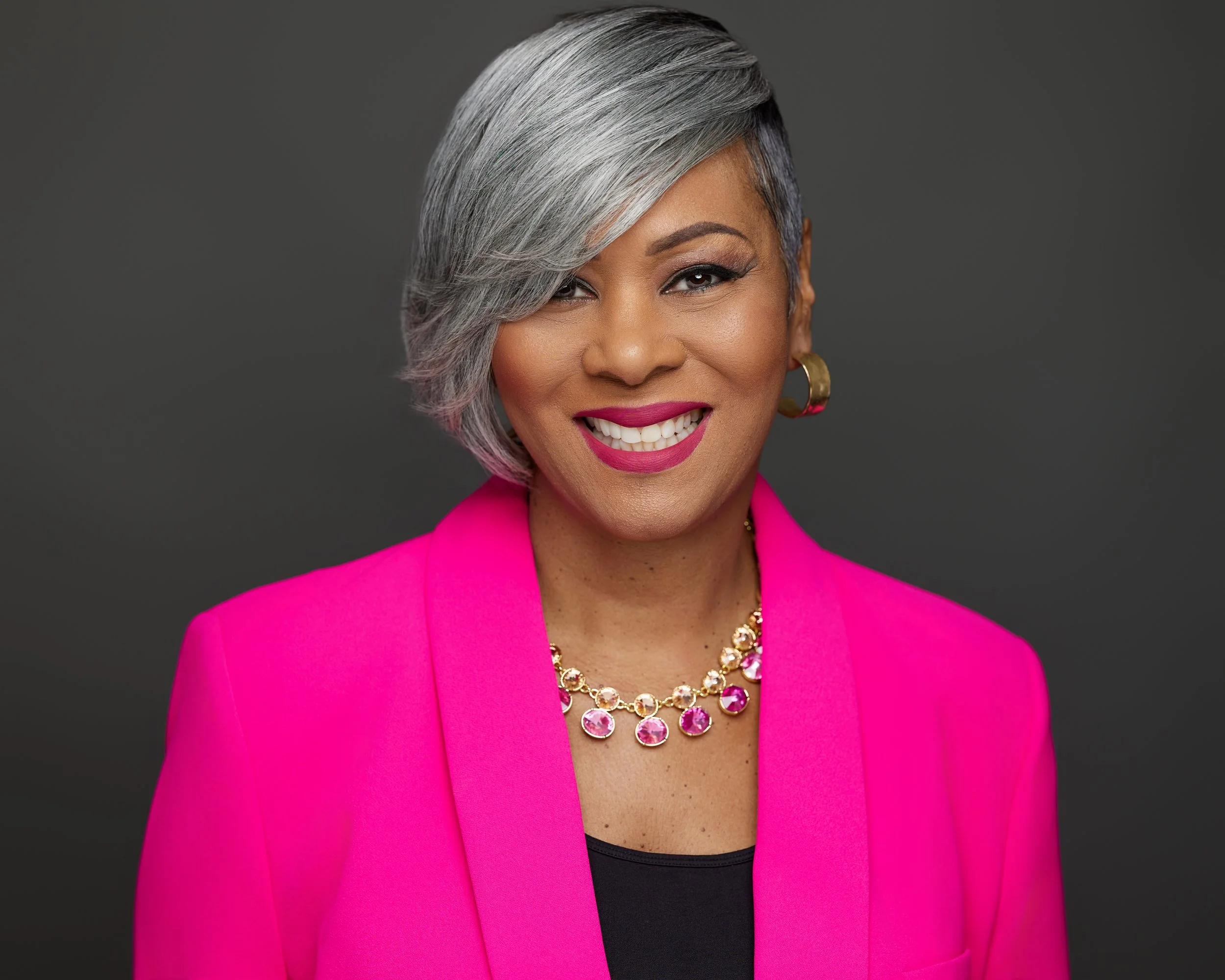 A woman with short, gray hair styled in a side-swept manner, smiling and wearing a bright pink blazer, black top, gold and pink jewelry, and earrings, against a dark gray background.