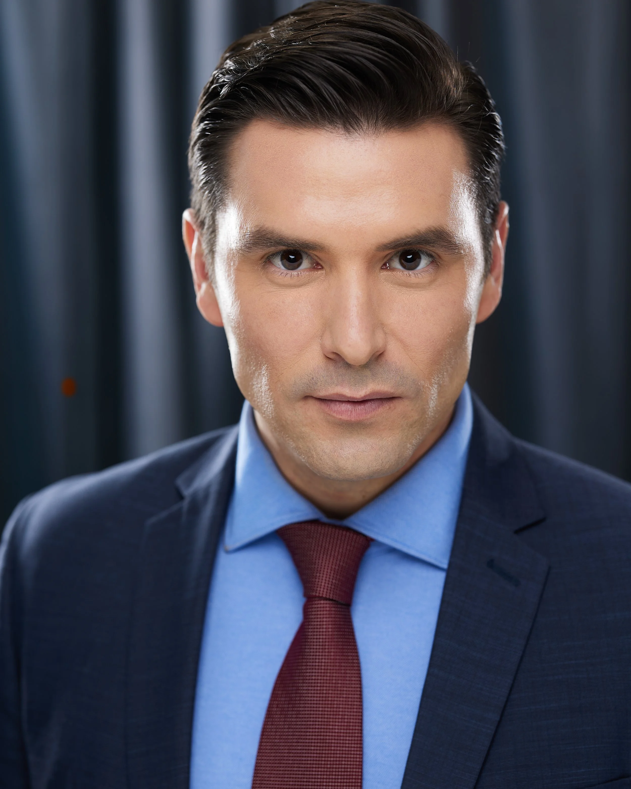 Portrait of a man in a navy suit, light blue shirt, and red tie, with dark hair styled back, standing in front of blue curtains.