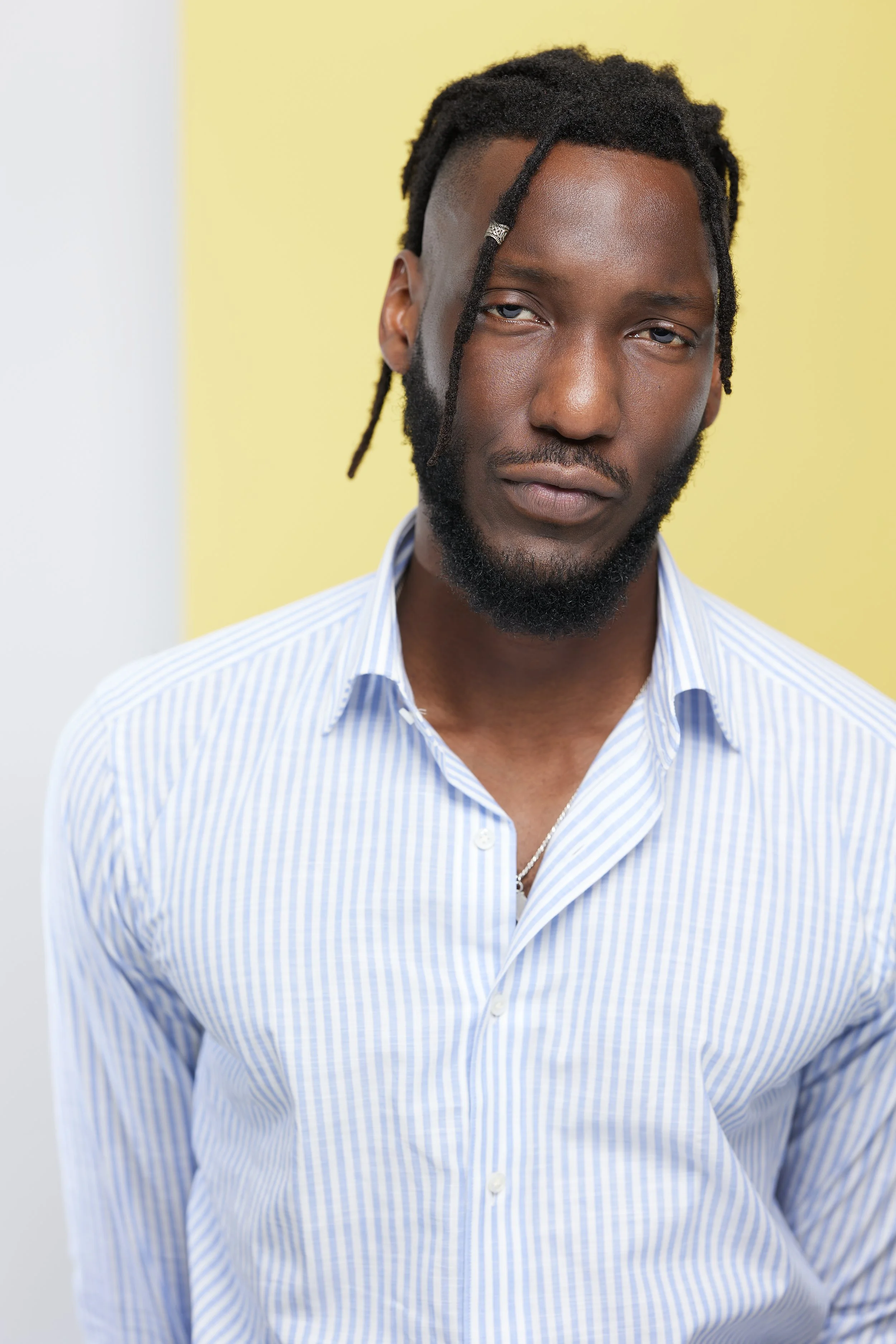A man with medium-dark skin tone, dreadlocks, and a beard wearing a blue and white striped button-up shirt, standing against a yellow background.