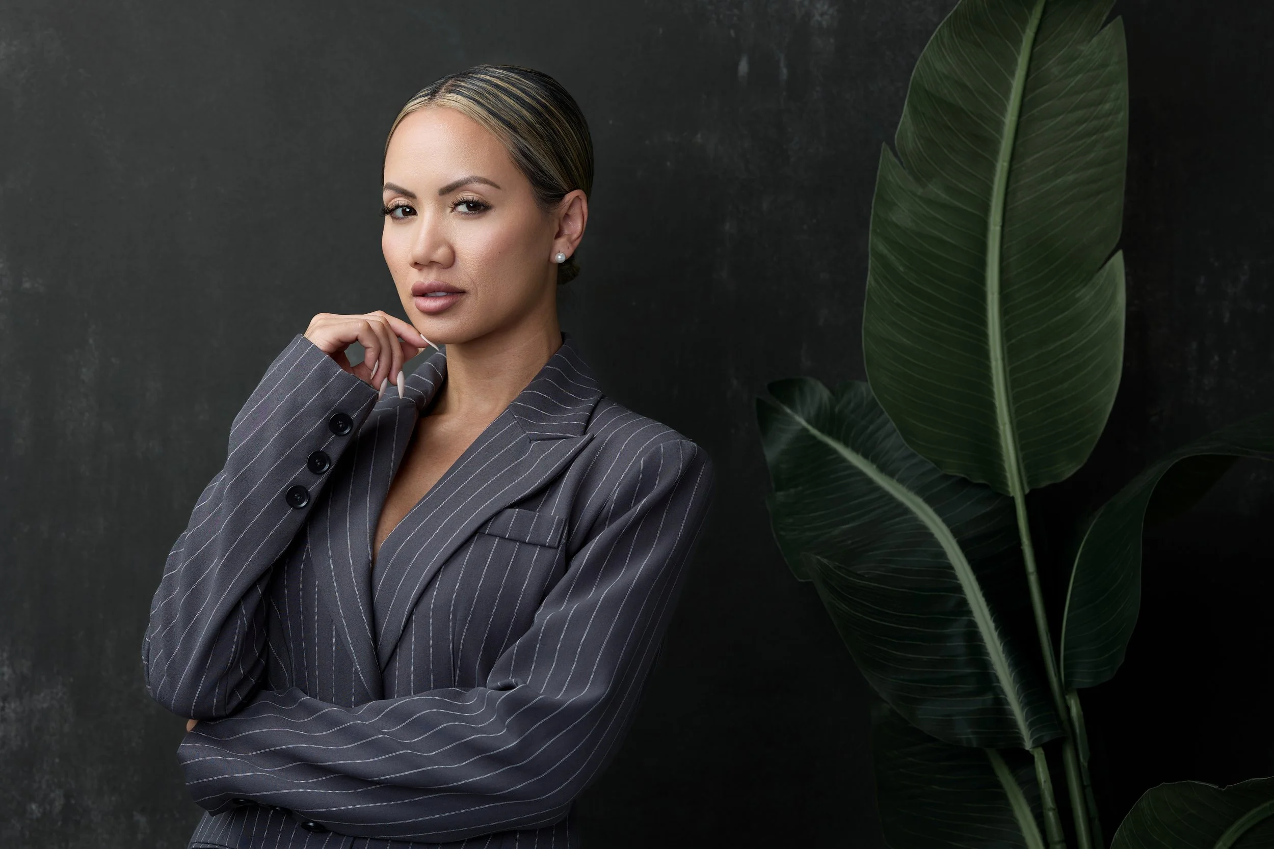 A confident woman in a gray pinstripe suit with blonde highlights posing against a dark background with large green leaves.