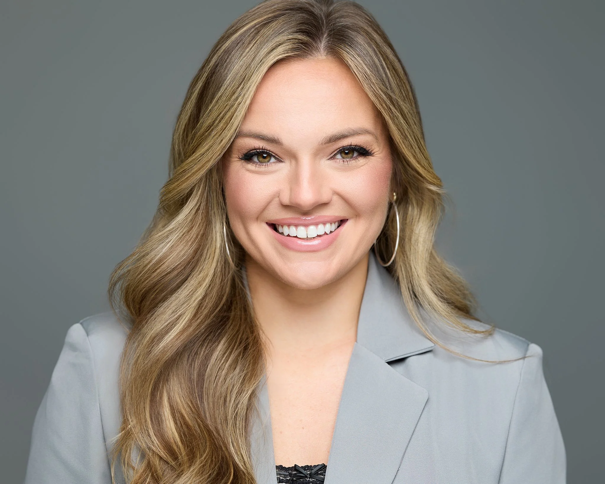 A woman with long, wavy blonde hair smiling against a gray background, wearing a light gray blazer and hoop earrings.
