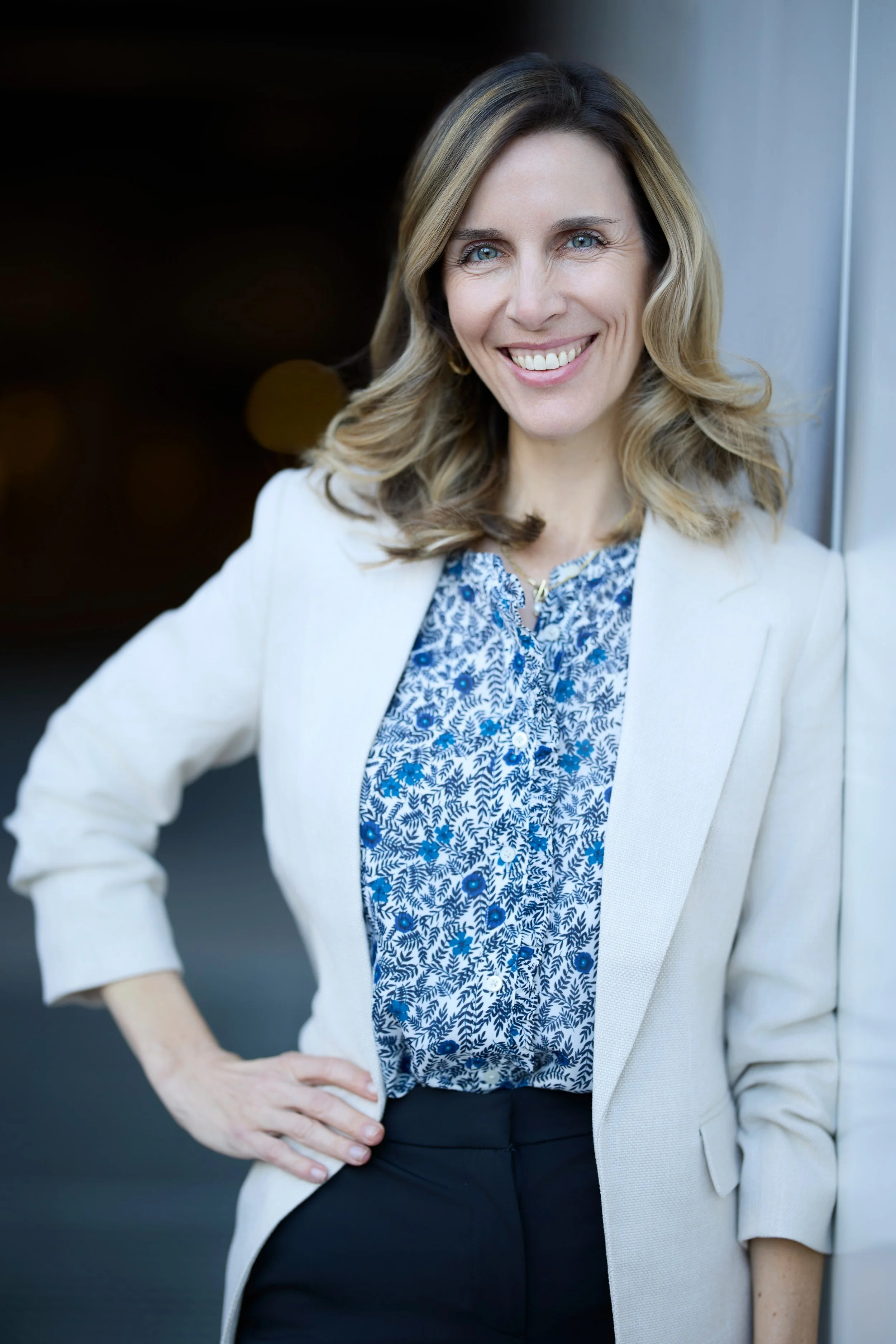 A woman with blonde, wavy hair and blue eyes smiling, wearing a white blazer over a blue patterned blouse, and black pants, standing outdoors near a wall.