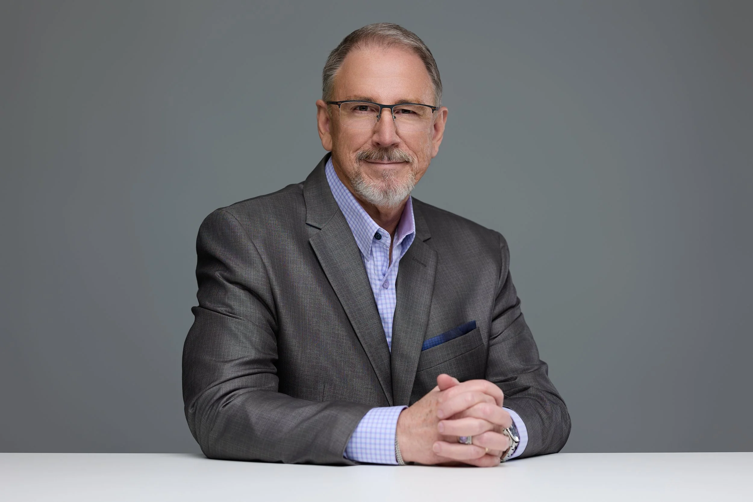 A mature man with glasses, a gray beard, wearing a gray suit and checkered shirt, sitting at a white table against a gray background, smiling with hands clasped.