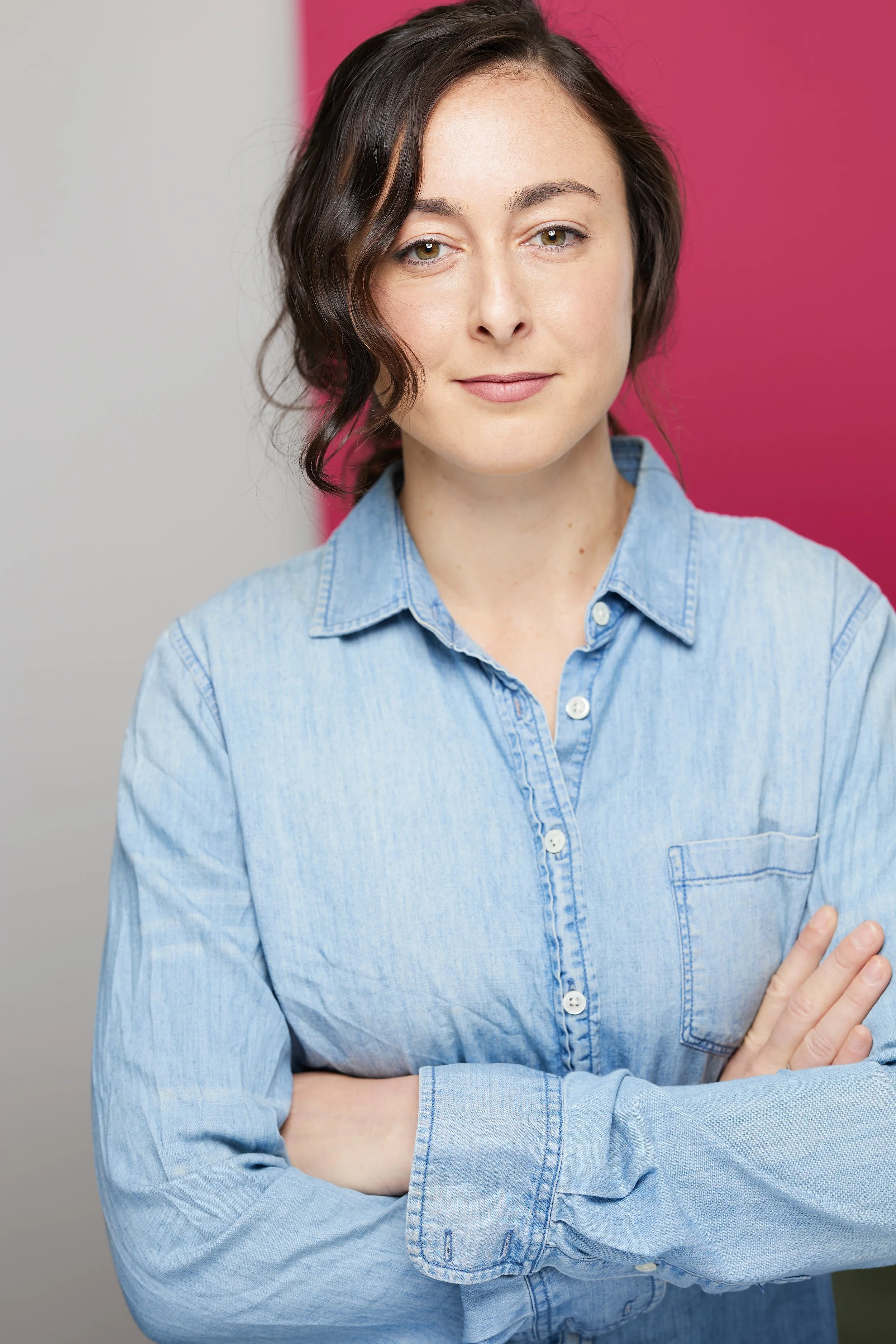 A woman with short dark hair and light skin, wearing a light blue denim shirt, standing with arms crossed in front of a background with pink and gray sections.