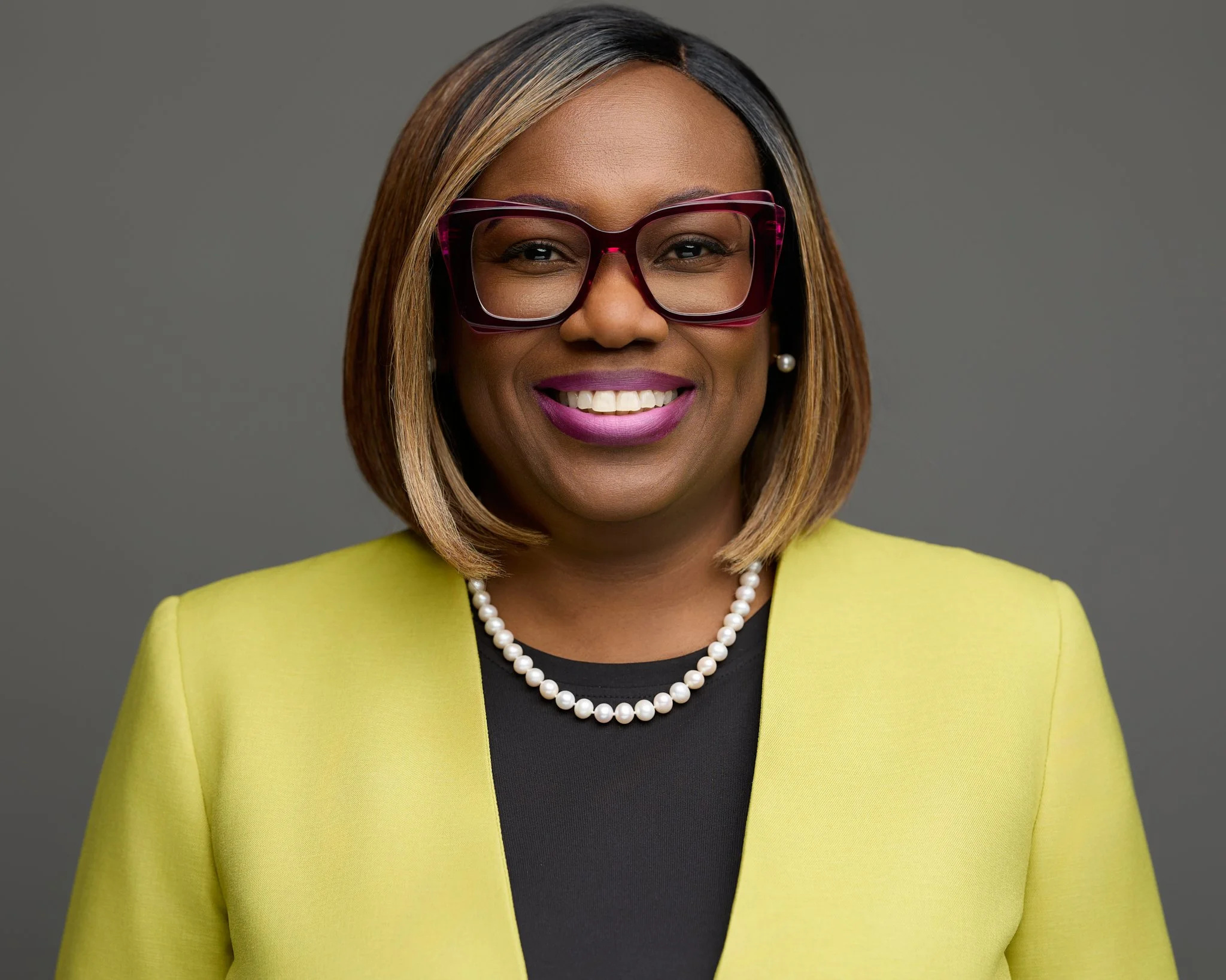 A woman with shoulder-length hair wearing glasses, a yellow blazer, a black top, pearl necklace, and pearl earrings, smiling against a gray background.