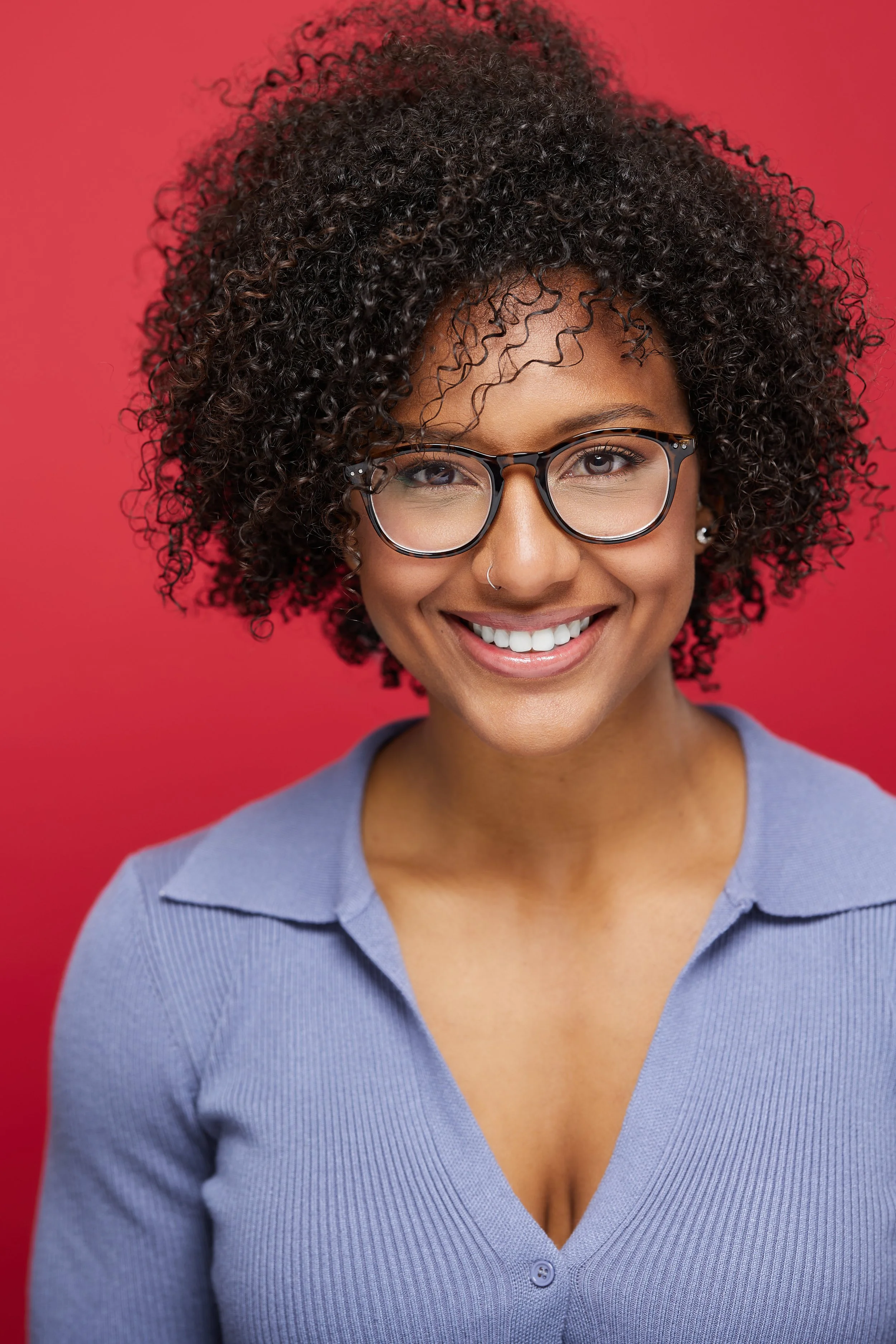 A woman with curly hair, glasses, and a nose ring smiling against a red background.